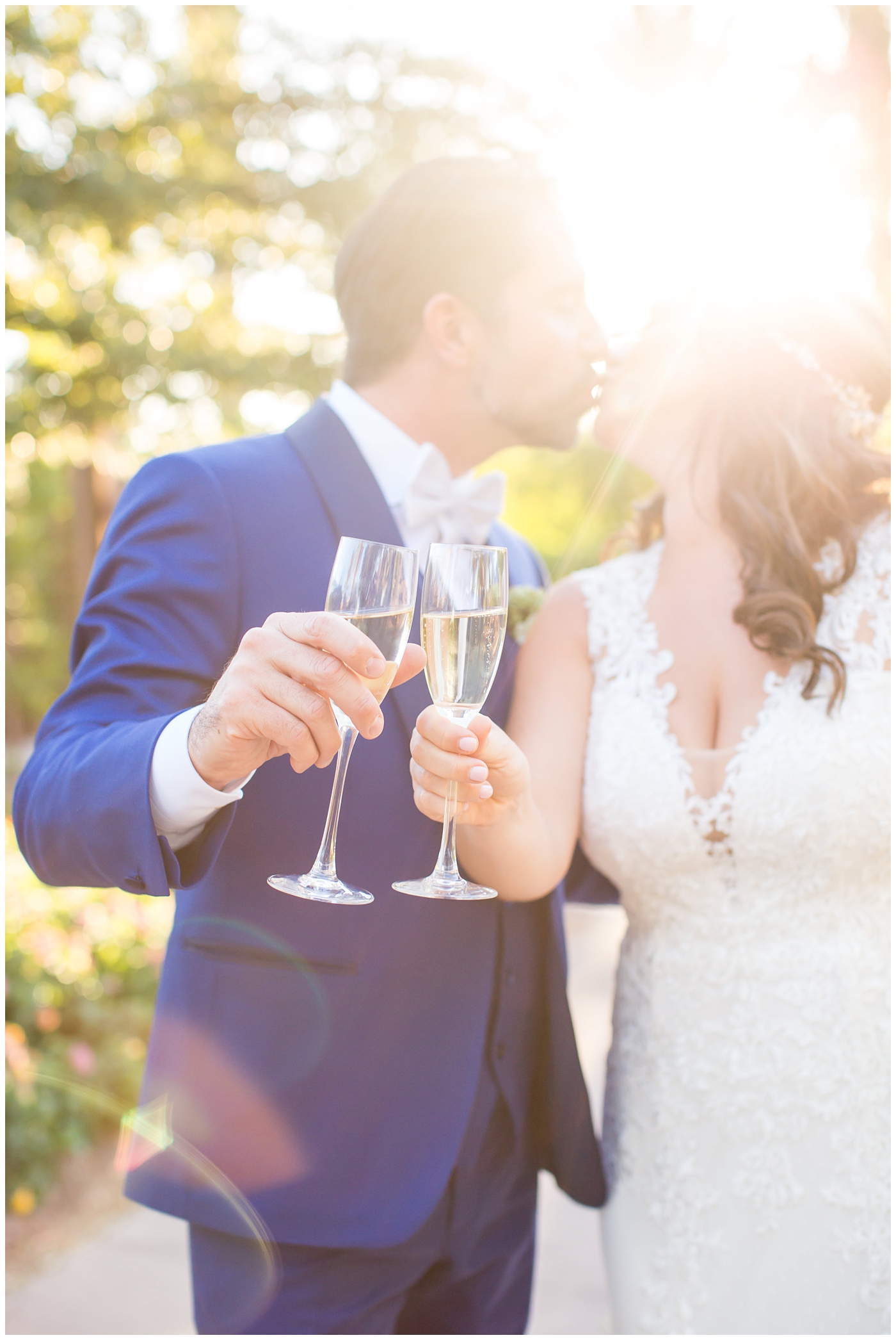 Bride in Justina Alexander dress with groom in custom Brother's Tailors blue suit kissing while holding champagne