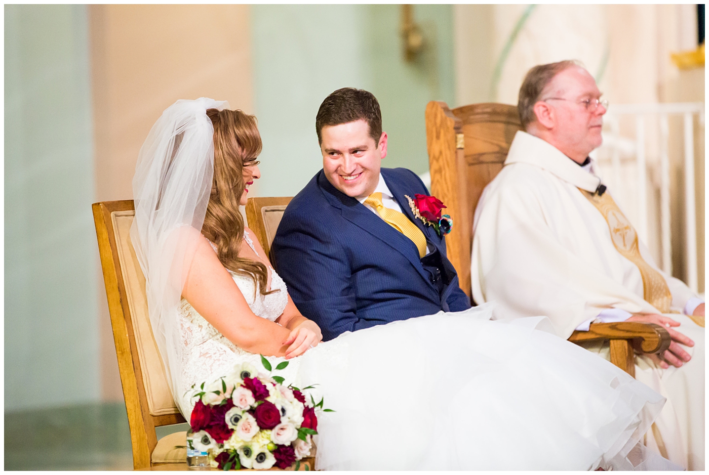 groom in blue suit with yellow tie and red rose boutonniere and bride in morilee wedding dress with red and white rose bouquet during ceremony