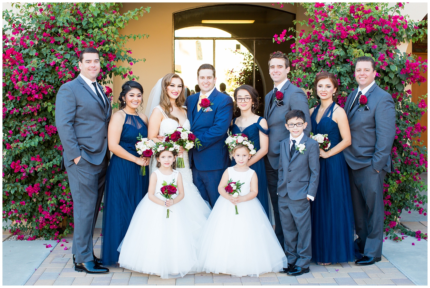 bridesmaids in blue dresses with bride in morilee wedding dress with red and white bouquet and groom in blue suit with yellow tie and red rose boutonniere and groomsmen in gray suits