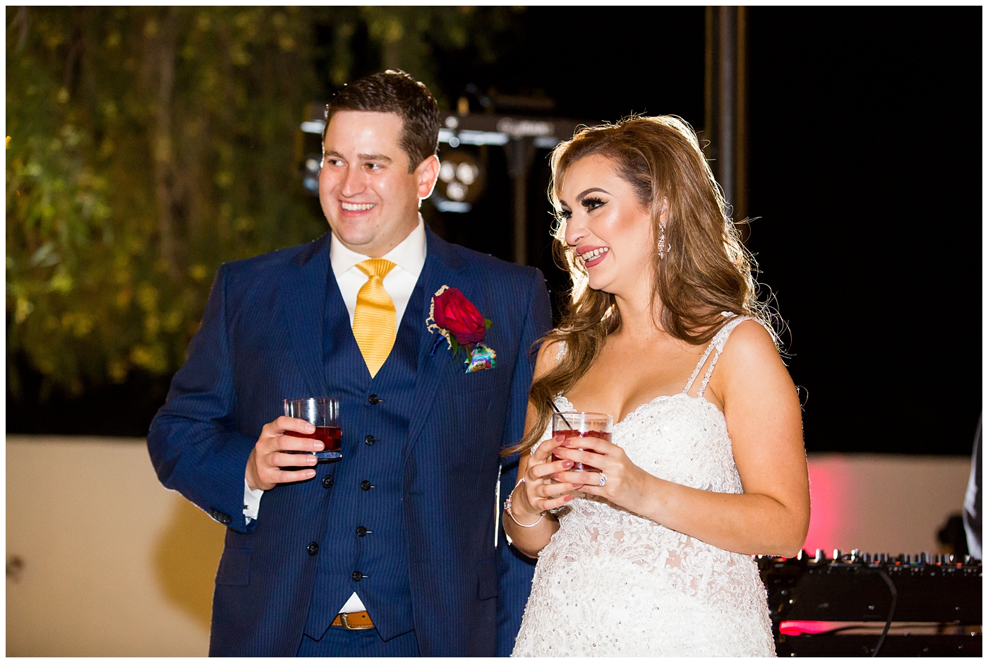 groom in blue suit with yellow tie and red rose boutonniere and bride in morilee wedding dress with red and white rose bouquet during toasts at reception