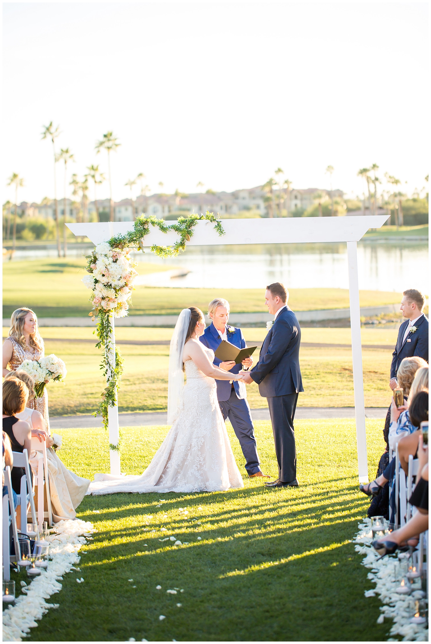bride in lillian lottie couture wedding dress with white rose bouquet and groom in navy blue suit from men's warehouse bride and groom during wedding ceremony on golf course