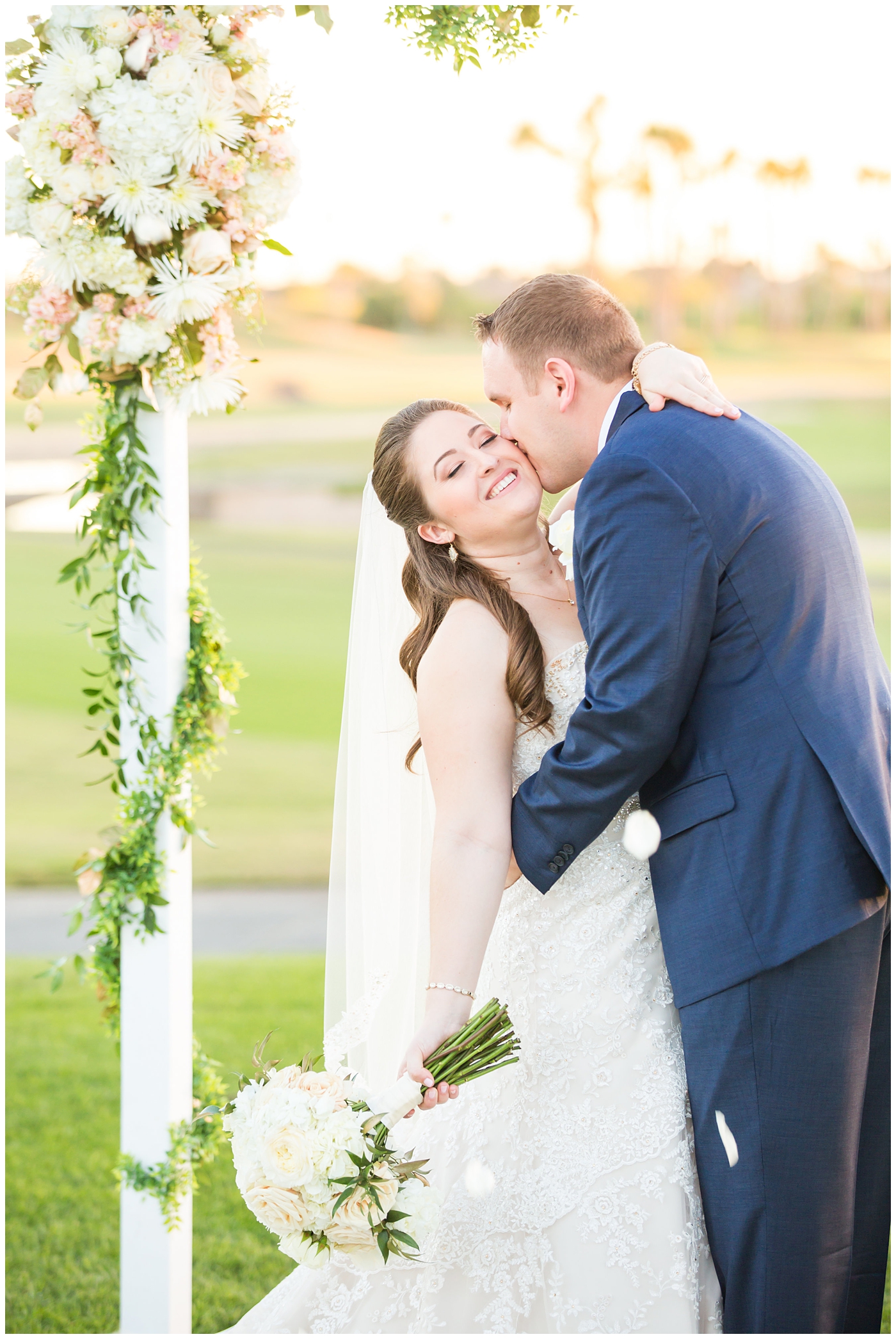 bride in lillian lottie couture wedding dress with white rose bouquet and groom in navy blue suit from men's warehouse bride and groom wedding portrait under arch on golf course