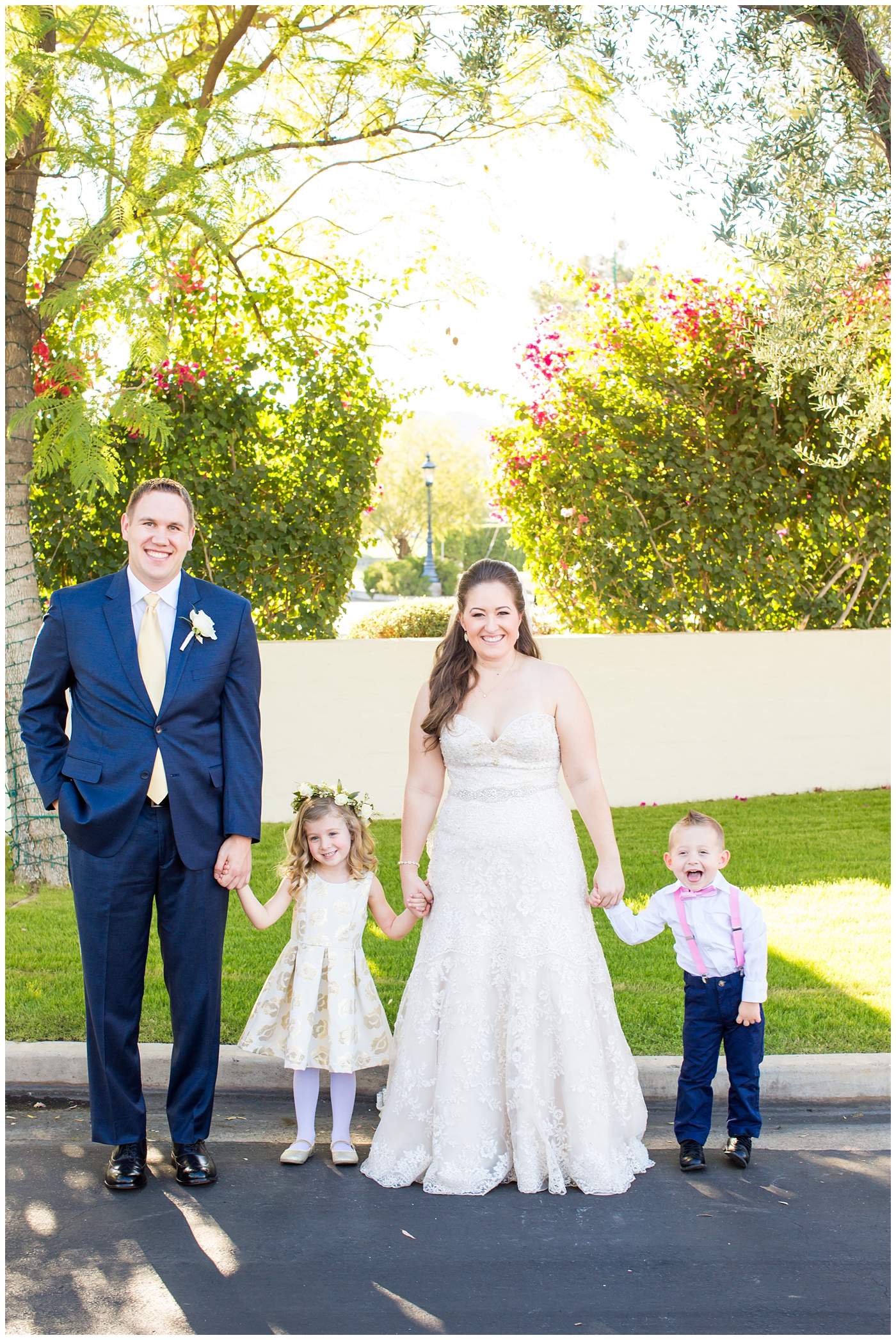 bride in lillian lottie couture wedding dress with white rose bouquet and groom in navy blue suit from men's warehouse bride and groom with flower girl wearing flower crown and ring bearer in suspenders