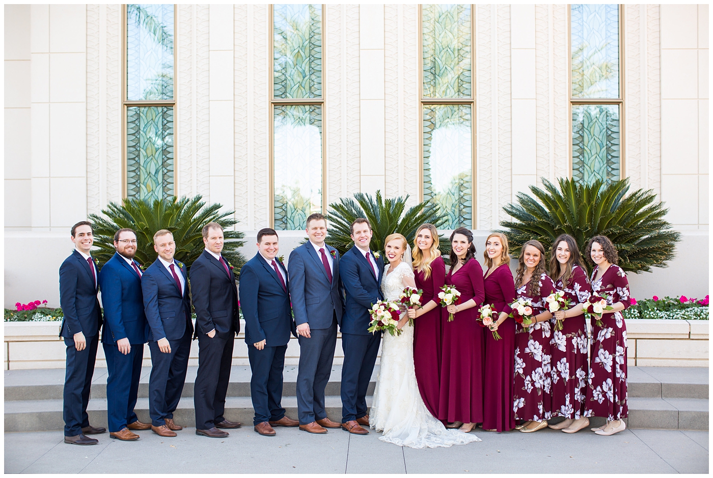 bride in sleeved lace dress with burgundy, white, and green wedding bouquet with bridesmaids in long sleeve burgundy dresses and groom in navy suit with burgundy tie with groomsmen wedding party portrait at Gilbert LDS temple