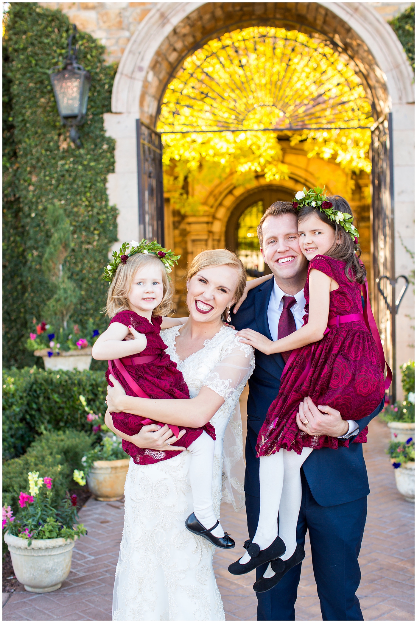 bride in sleeved lace dress with burgundy, white, and green wedding bouquet and groom in navy suit with burgundy tie wedding day portrait with flower girls with floral head bands and burgundy dresses at Villa Siena