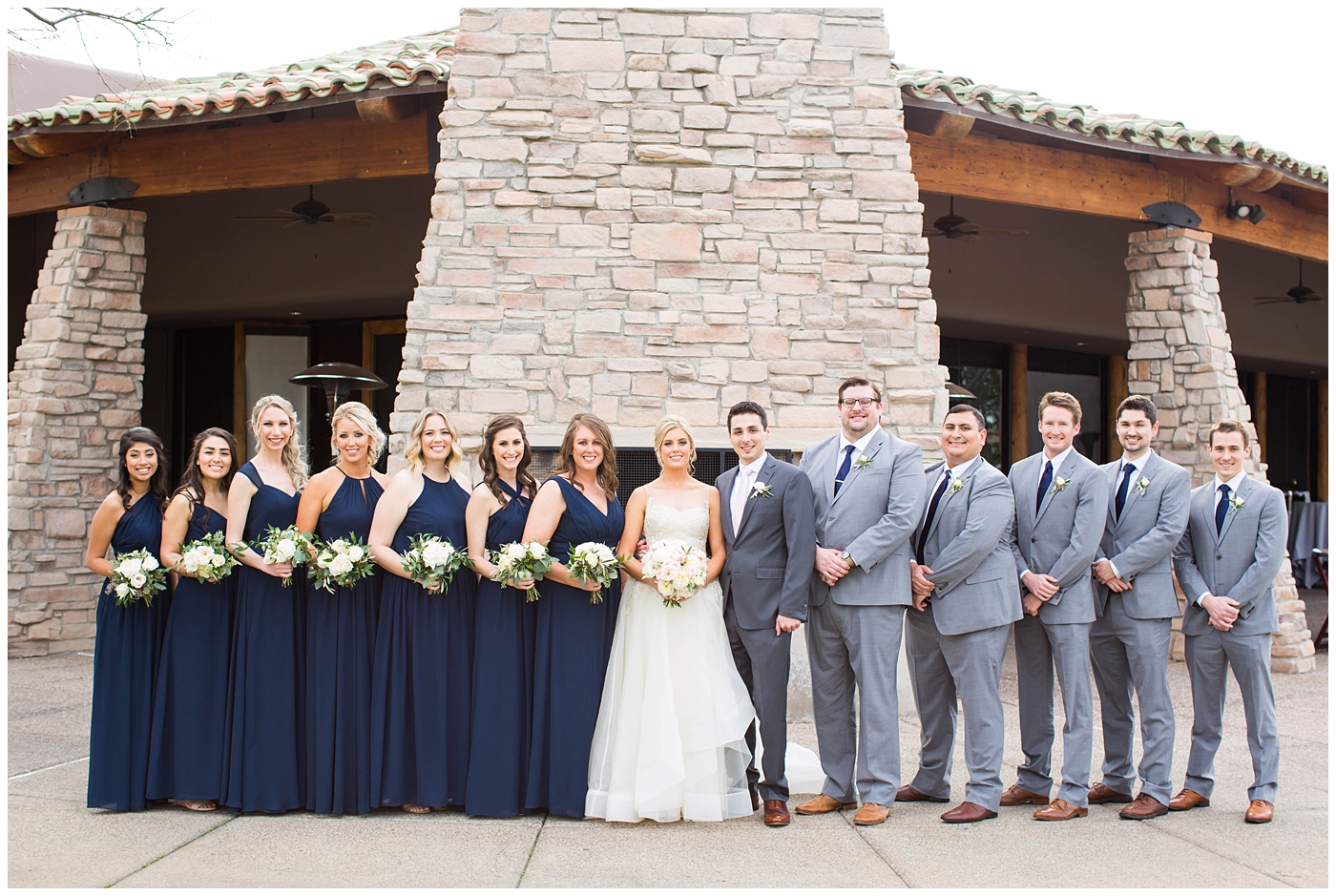 bridesmaids in assorted navy blue dresses with white rose and greenery flower bouquets with groom in gray suit with pink tie with groomsmen with navy ties on wedding day bridal party picture