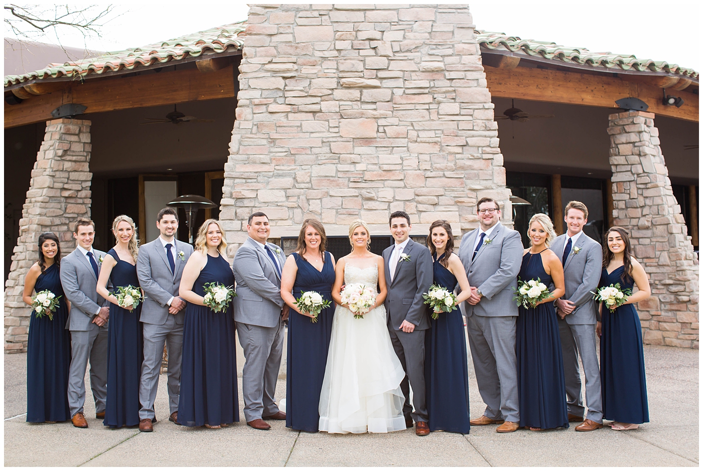 bridesmaids in assorted navy blue dresses with white rose and greenery flower bouquets with groom in gray suit with pink tie with groomsmen with navy ties on wedding day bridal party picture