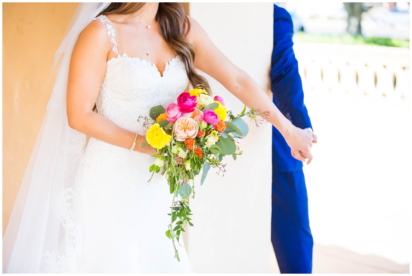 bride in lace cap sleeve wedding dress with bright pink, yellow and green flower bouquets first touch with groom in blue suit on wedding day at church
