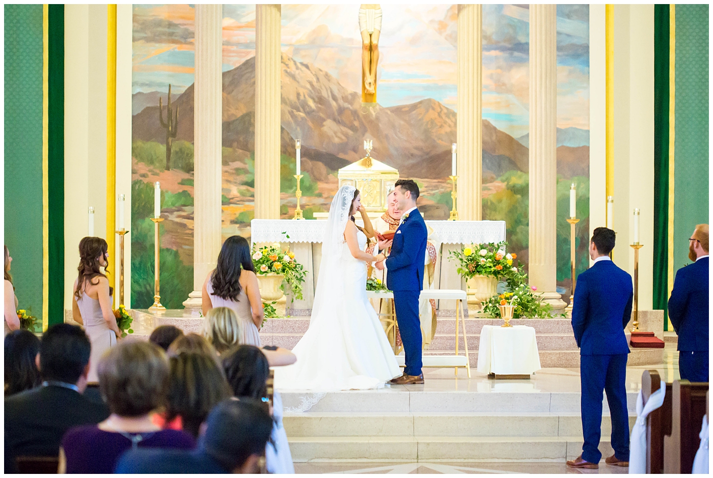 bride in lace cap sleeve wedding dress with bright pink, yellow and green flower bouquets with groom in blue suit on wedding day at church ceremony