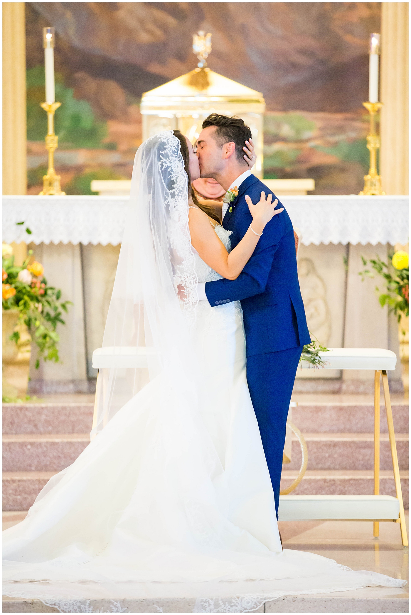 bride in lace cap sleeve wedding dress with bright pink, yellow and green flower bouquets with groom in blue suit on wedding day at church ceremony first kiss