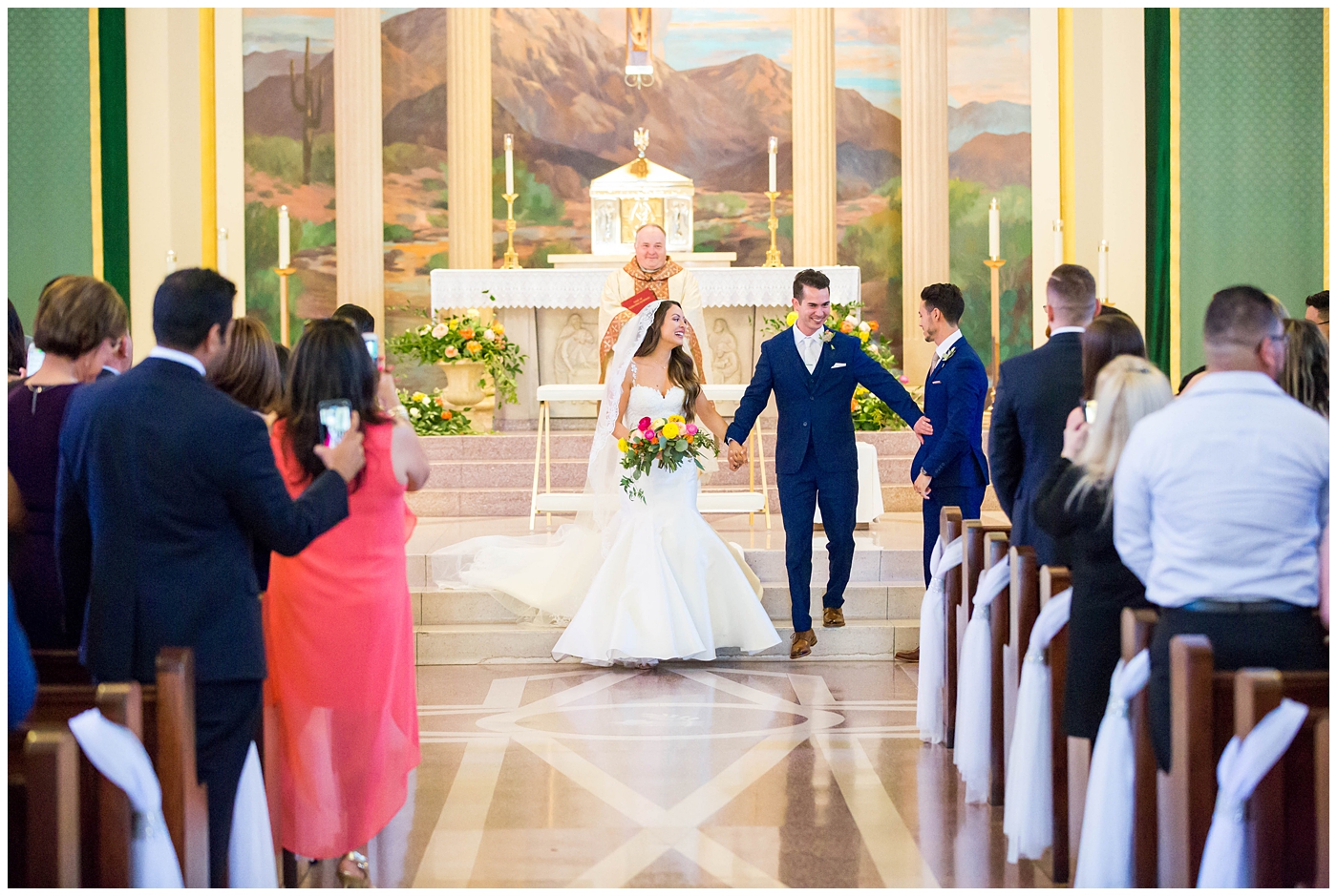 bride in lace cap sleeve wedding dress with bright pink, yellow and green flower bouquets with groom in blue suit on wedding day at church ceremony
