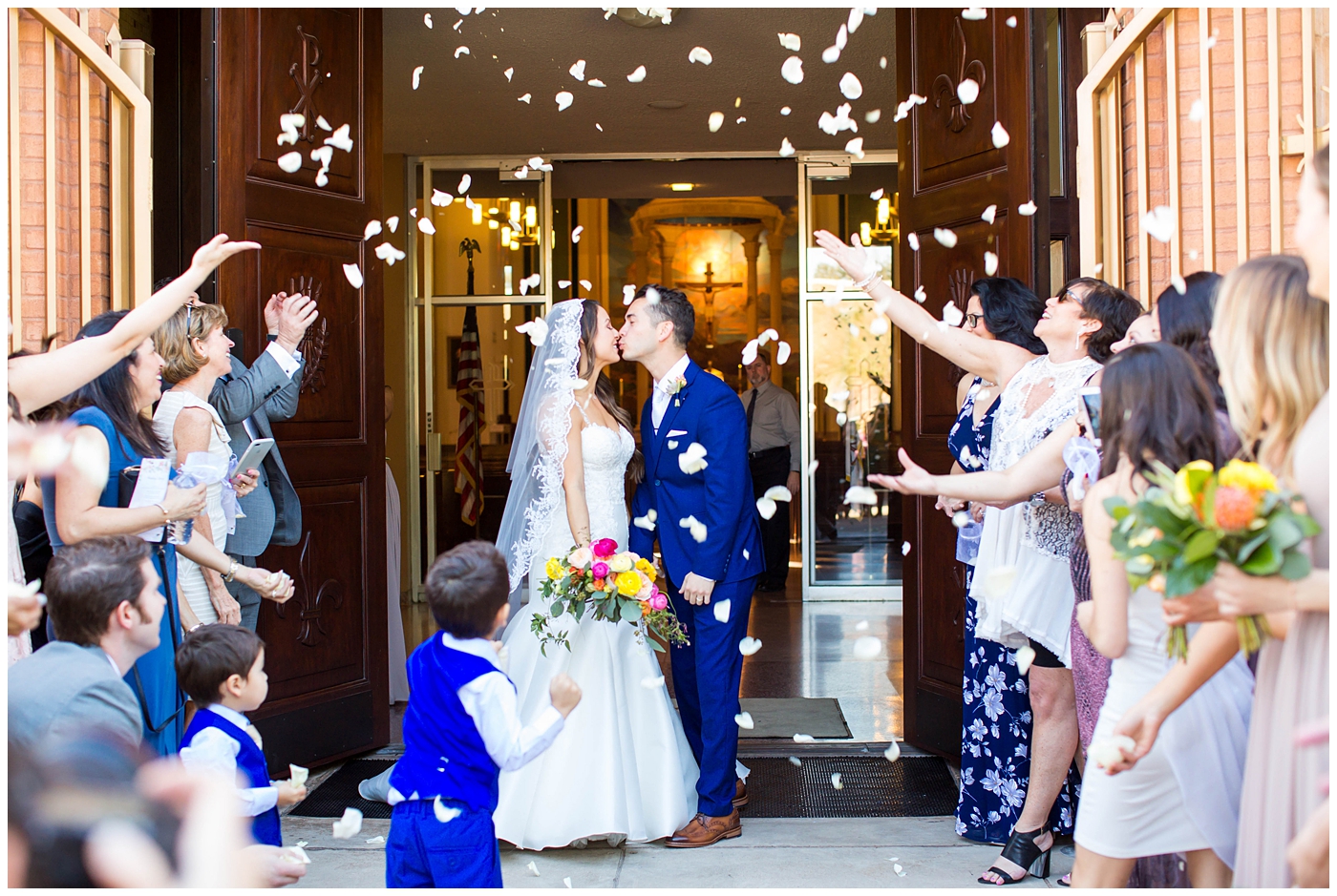 bride in lace cap sleeve wedding dress with bright pink, yellow and green flower bouquets with groom in blue suit on wedding day at church getting flower petals thrown as they exit