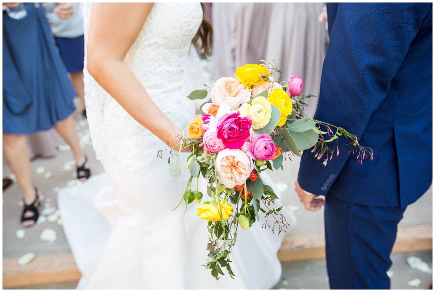 bride in lace cap sleeve wedding dress with bright pink, yellow and green flower bouquets fwith groom in blue suit on wedding day portrait at church
