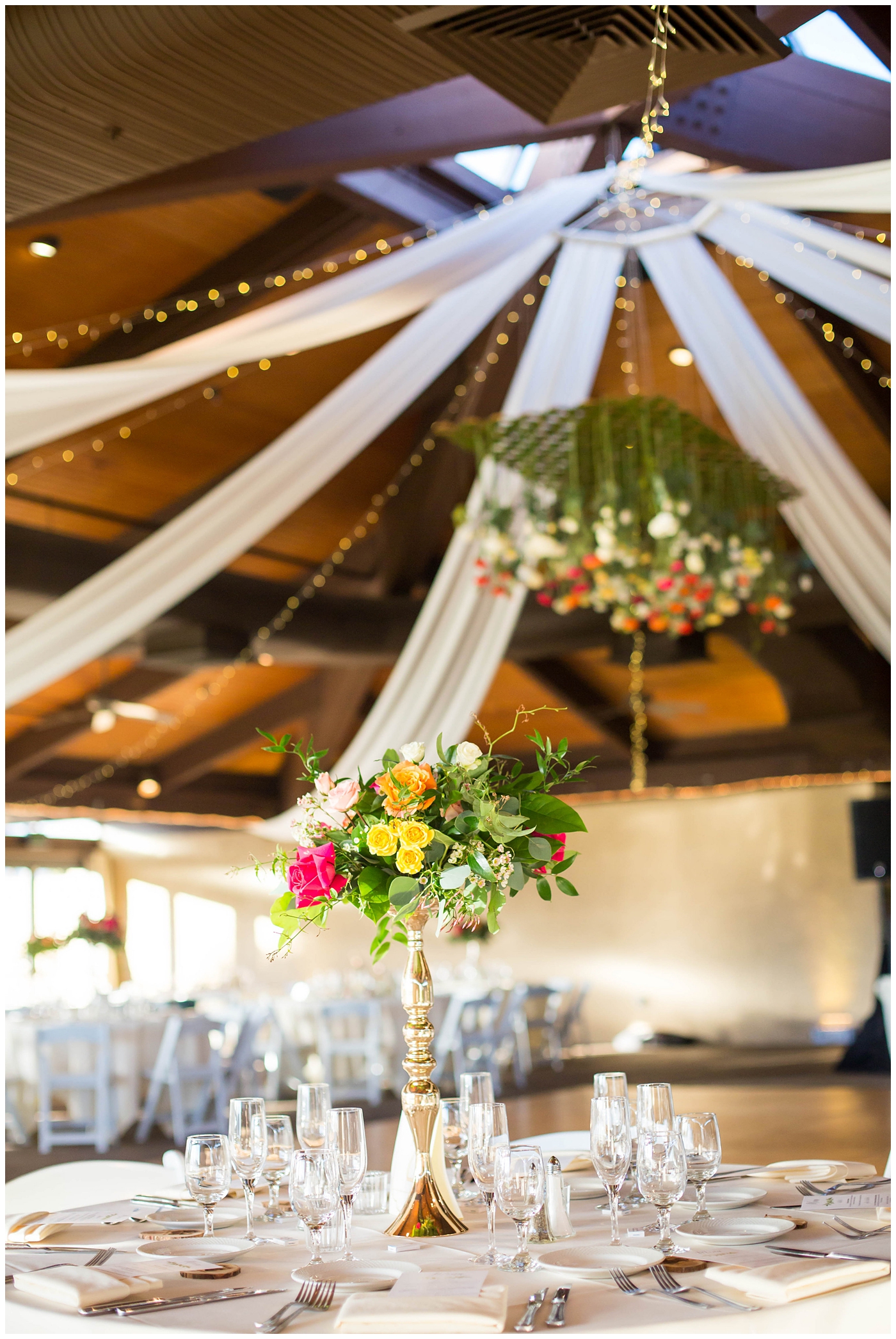 wedding reception details with white tables and chairs with gold stands with white, bright pink, orange, yellow and green flowers hanging from ceiling