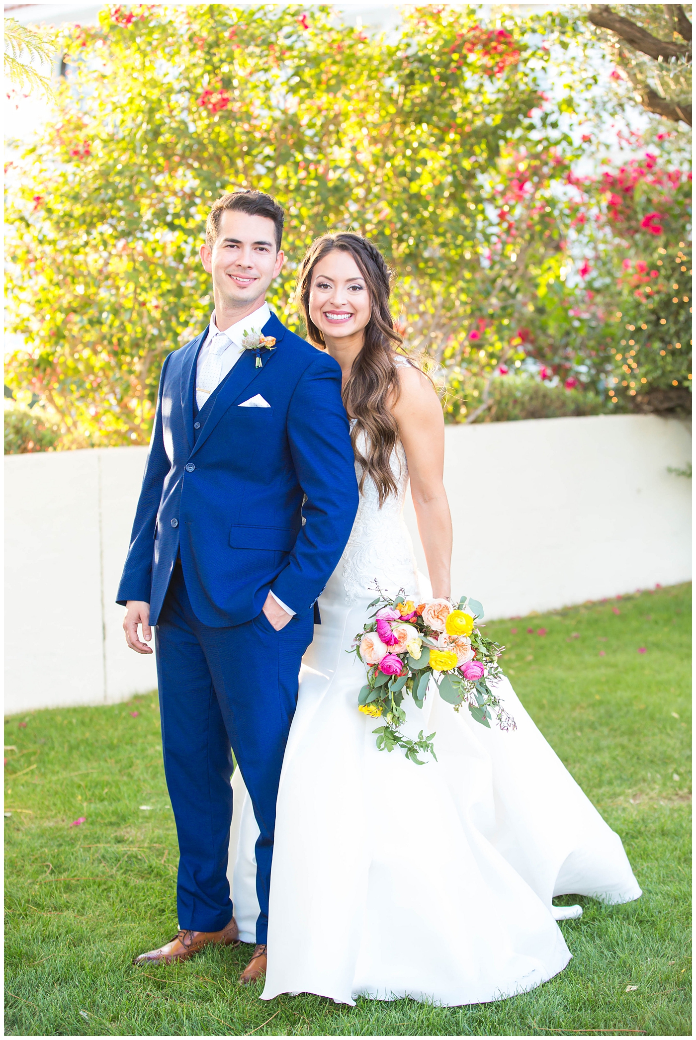 bride in lace cap sleeve wedding dress with bright pink, yellow and green flower bouquets and flower crown with groom in blue suit on wedding day portrait 