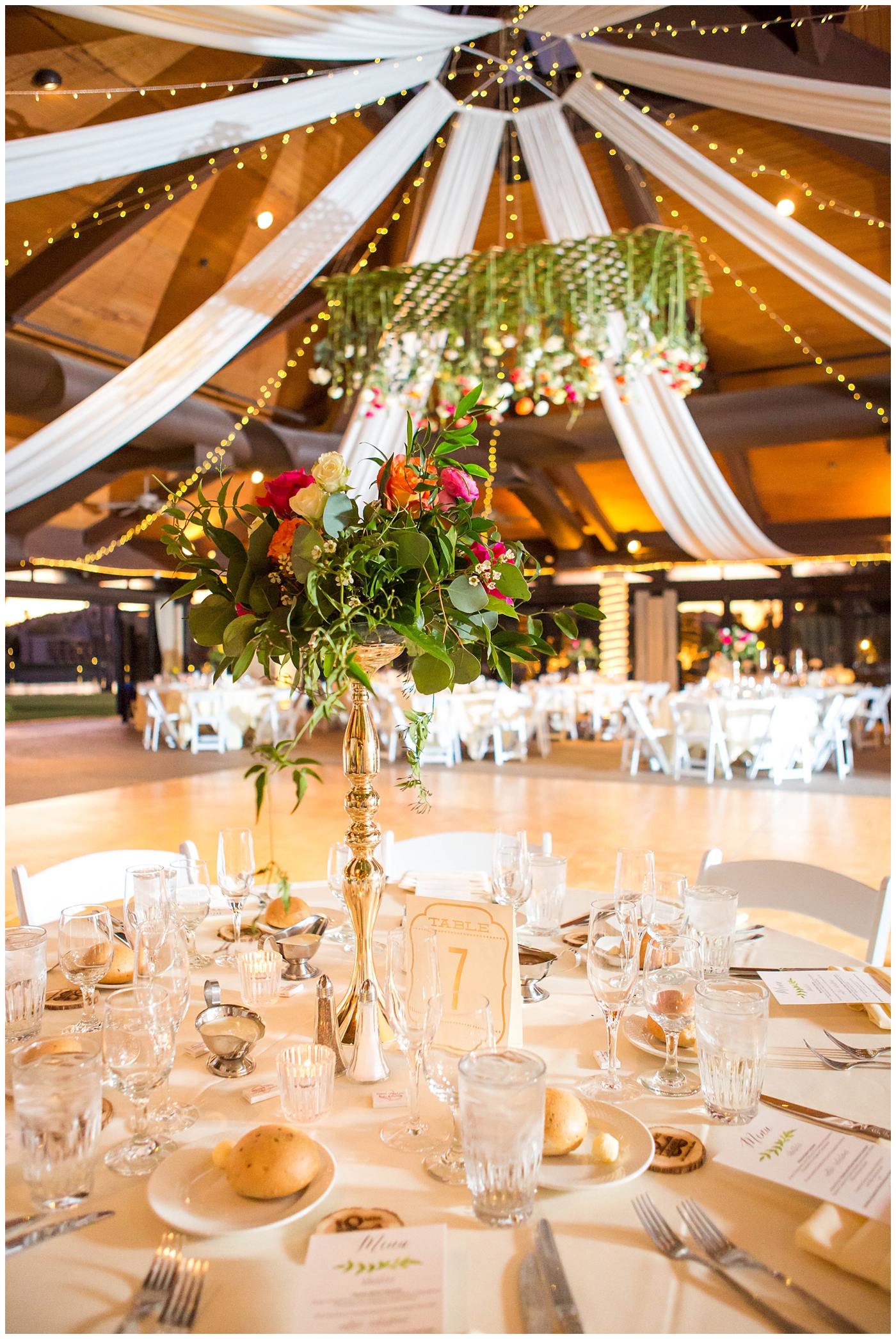 wedding reception details with white tables and chairs with gold stands with white, bright pink, orange, yellow and green flowers hanging from ceiling