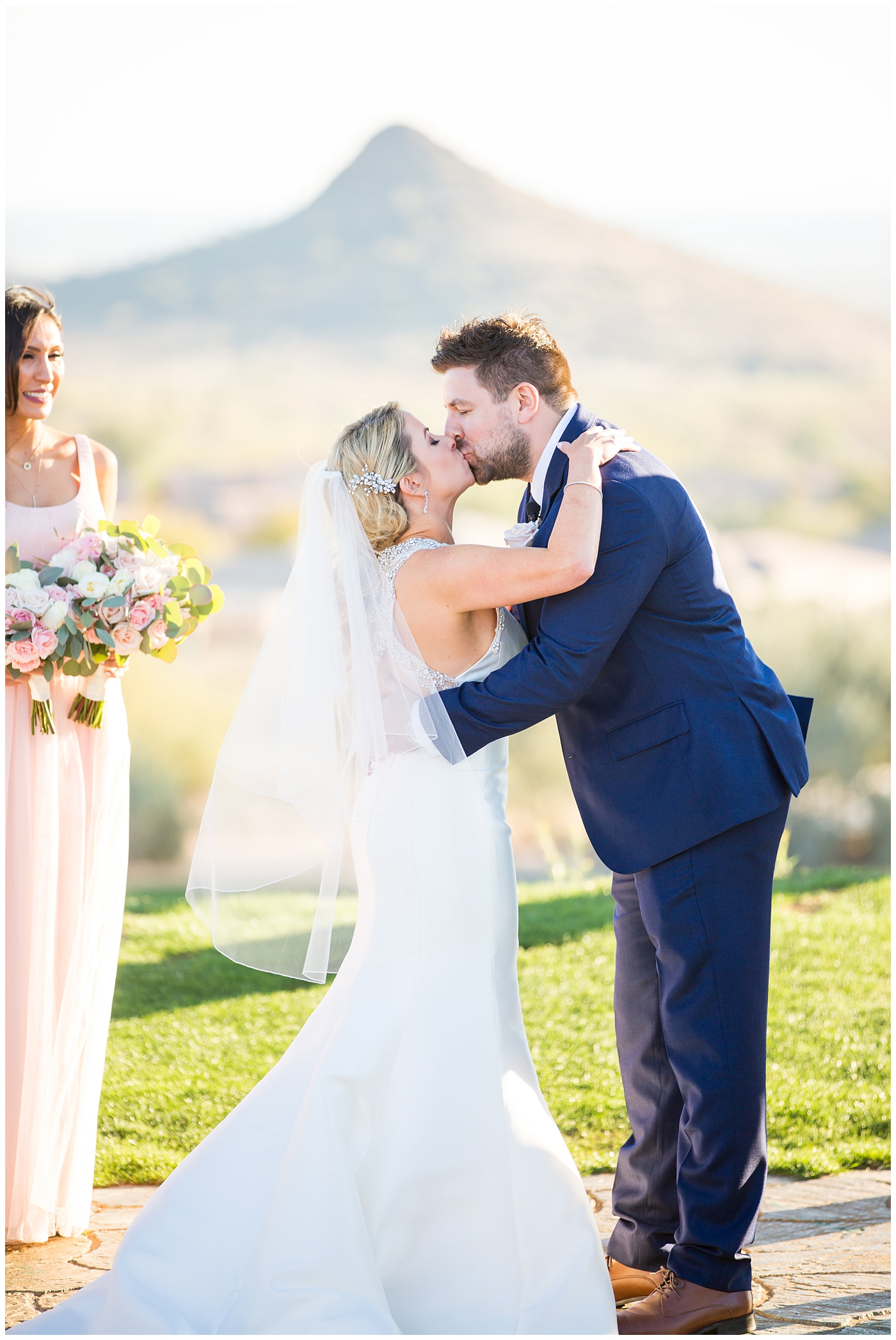 gorgeous bride with side swept hair in racerback pronovias dress and blush pink, white rose and eucalyptus greenery wedding bouquet with groom in navy suit and tie first kiss at wedding ceremony outside