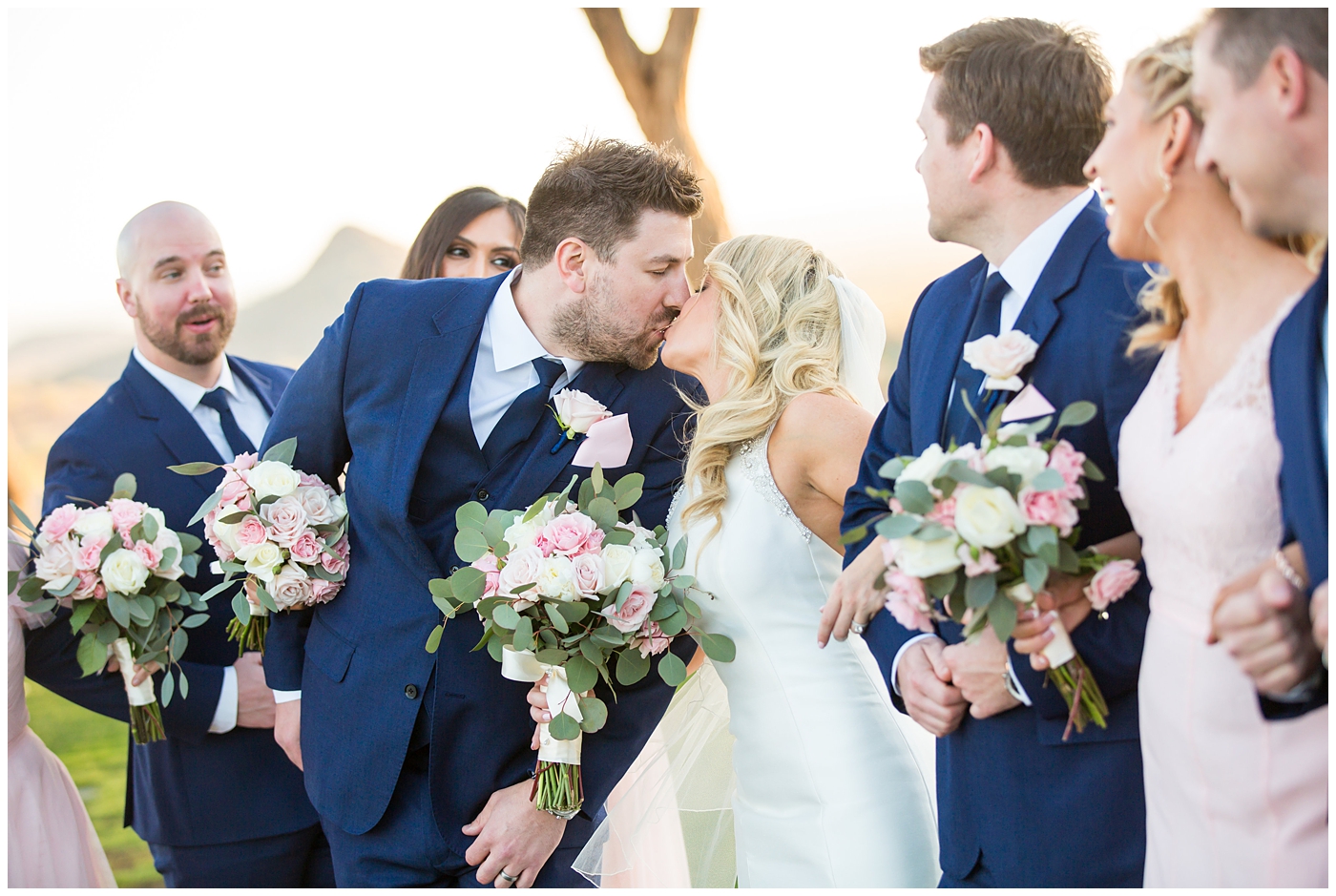 gorgeous bride with side swept hair in racerback pronovias dress and blush pink, white rose and eucalyptus greenery wedding bouquet with groom in navy suit and tie with bridal party bridesmaids in blush pink dresses and groomsmen in navy blue suits on wedding day
