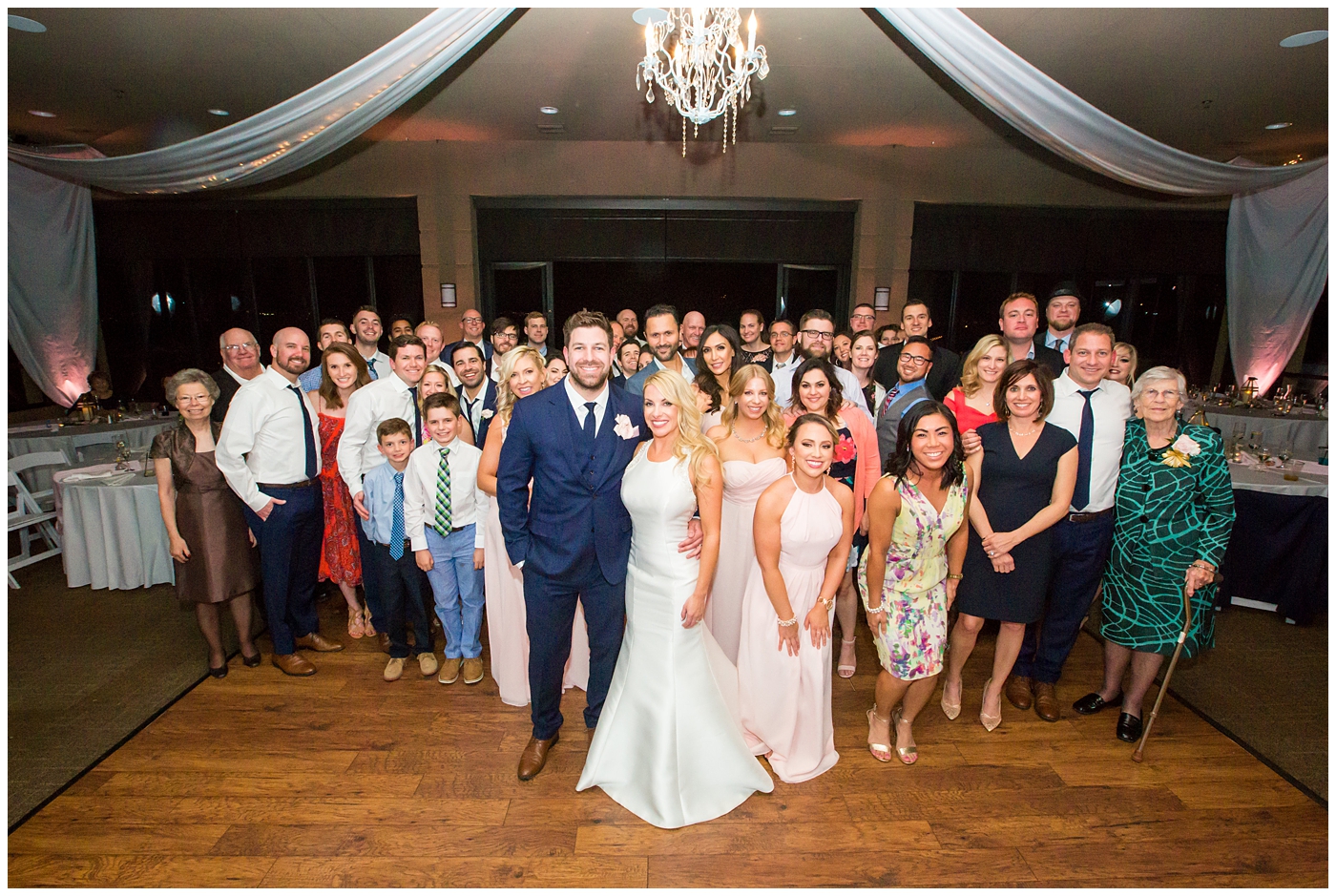 gorgeous bride with side swept hair in racerback pronovias dress and blush pink, white rose and eucalyptus greenery wedding bouquet with groom in navy suit and tie with all their guests at wedding reception in ballroom