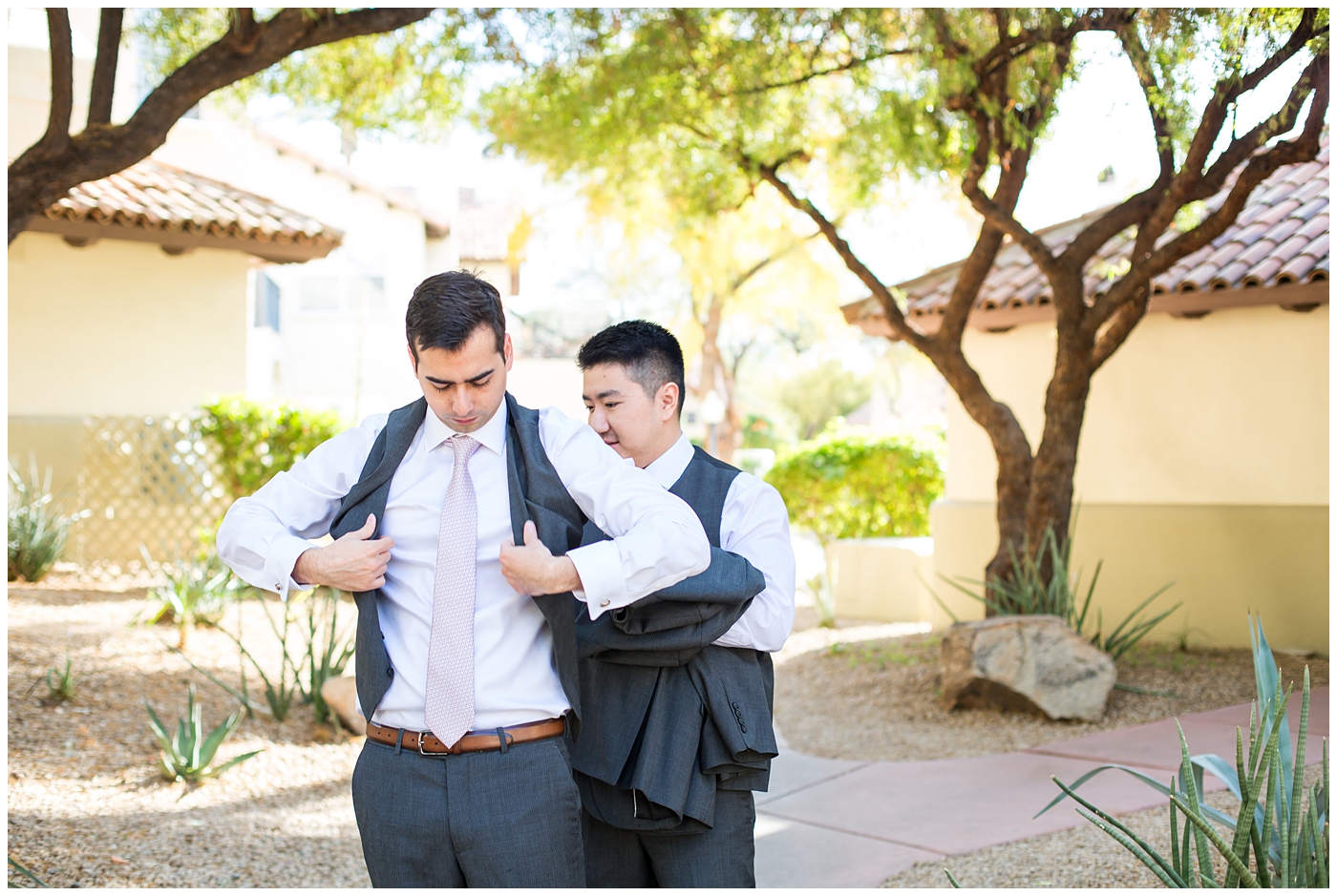 groom getting in gray suit with silver tie with help from best man on wedding day