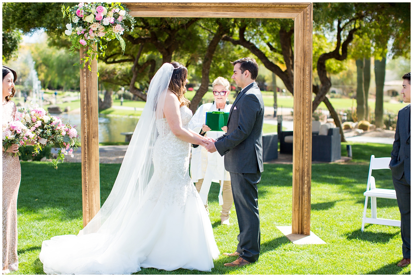 bride in strapless wedding dress with beads and shiny sparkle tool holding pink peony, roses, light coral rose and loose greenery bridal bouquet with groom in gray suit with silver tie standing under picture frame arch during outdoor wedding ceremony at fairmont princess