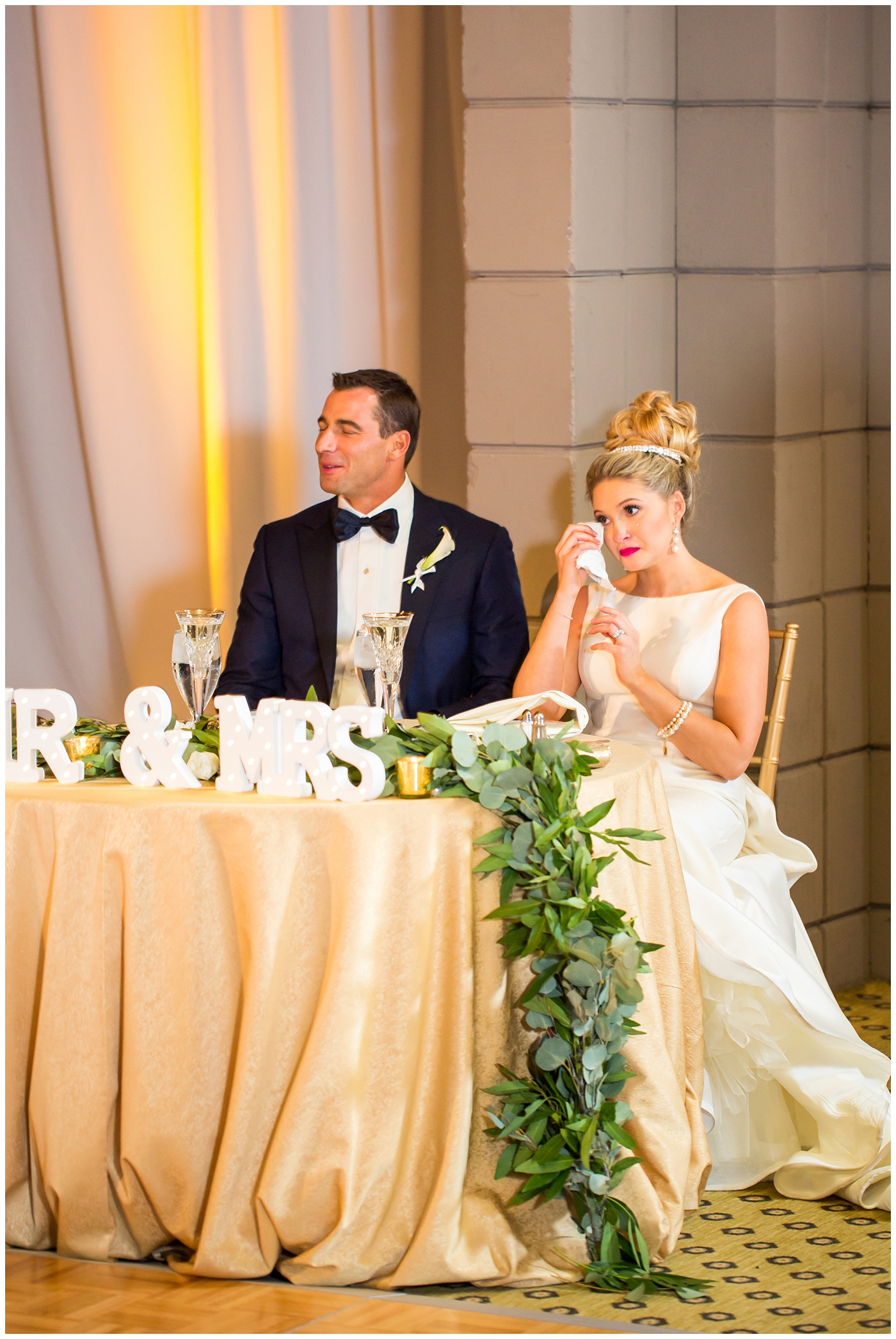 Bride in sareh nouri wedding dress with bow on the back and pink lip stick with all white flower bouquet with groom in navy neiman marcus and canali suit with bowtie with calla lilly boutonniere wedding day ballroom reception during toasts