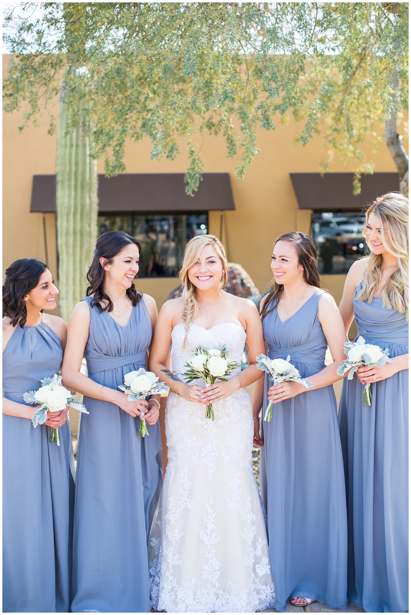 bride with fishtail braid and strapless dress with white rose and greenery bouquet with bridesmaids in slate gray long dresses getting ready on wedding day