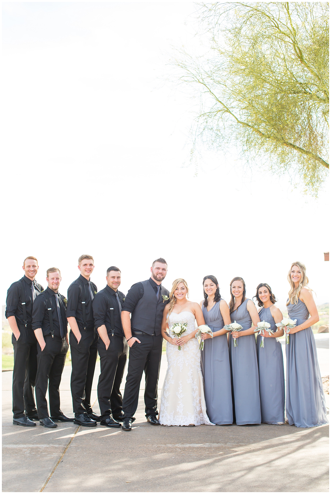 bride with fishtail braid and strapless dress with white rose and greenery bouquet with bridesmaids in slate gray long dresses with groom in black pants and vest and tie with rolled up shirt sleeves with succulent boutonniere with groomsmen in black pants with suspenders and tie wedding day bridal party portrait