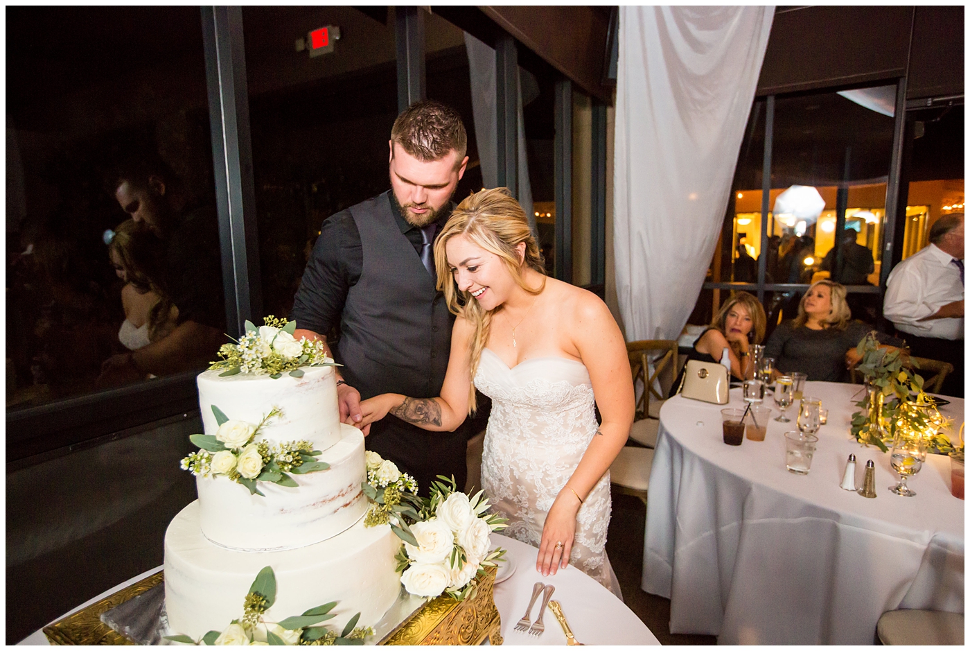 bride with fishtail braid and strapless dress with white rose and greenery bouquet with groom in black pants and vest and tie with rolled up shirt sleeves with succulent boutonniere wedding day portrait at reception cake cutting