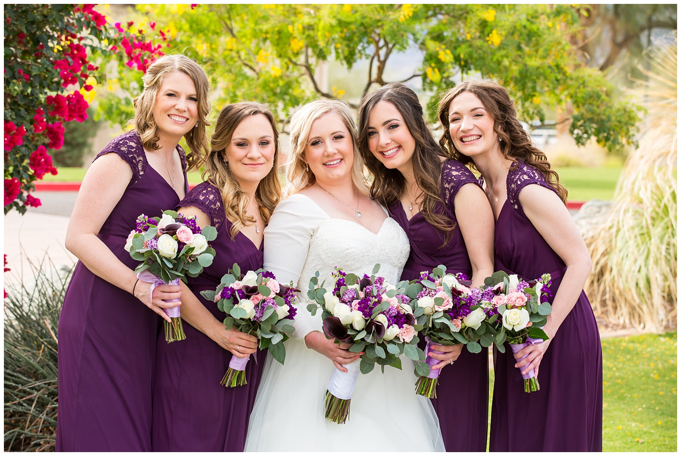 blonde bride in long sleeve wedding dress with purple and white flower bouquet with bridesmaids in plum dresses on wedding day bridal party portrait