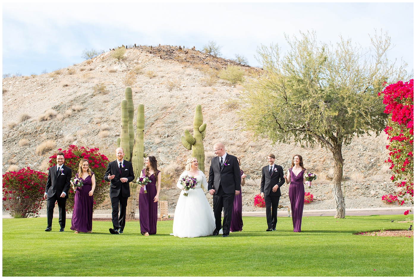 blonde bride in long sleeve wedding dress with purple and white flower bouquet with bridesmaids in plum dresses with groom in black suit with purple calla lilly boutonniere with groomsmen in black suits with plum vests on wedding day bridal party portrait