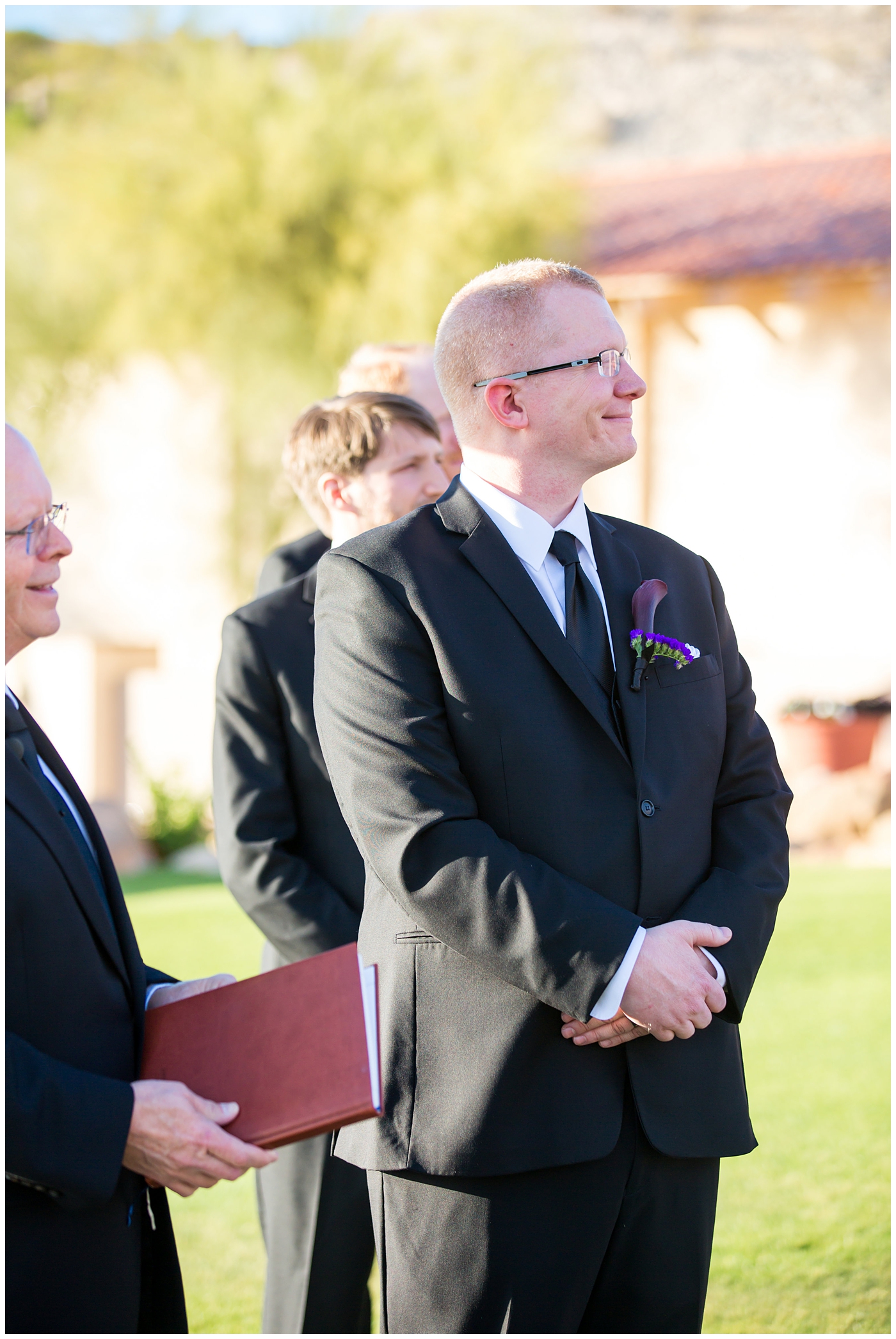 groom in black suit with purple calla lilly boutonniere waiting for bride to walk down the aisle at wedding ceremony