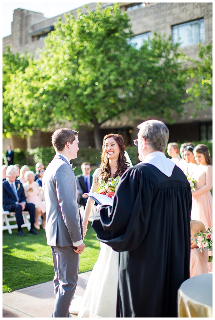 bride in matthew christopher wedding dress with cap sleeves with white, pink and orange ranunculus flowers and greenery bouquet with groom in light gray suit with tie wedding day ceremony at Arizona Biltmore