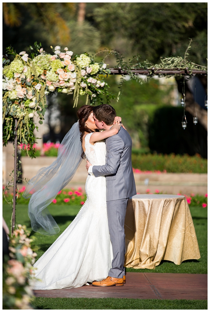 bride in matthew christopher wedding dress with cap sleeves with white, pink and orange ranunculus flowers and greenery bouquet with groom in light gray suit with tie wedding day ceremony first kiss at Arizona Biltmore
