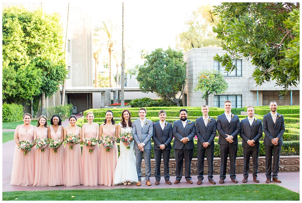 bride in matthew christopher wedding dress with cap sleeves with white, pink and orange ranunculus flowers and greenery bouquet with groom in light gray suit with tie with wedding party bridesmaids in blush dresses and groomsmen in dark gray suits wedding day portraits in Arizona biltmore garden