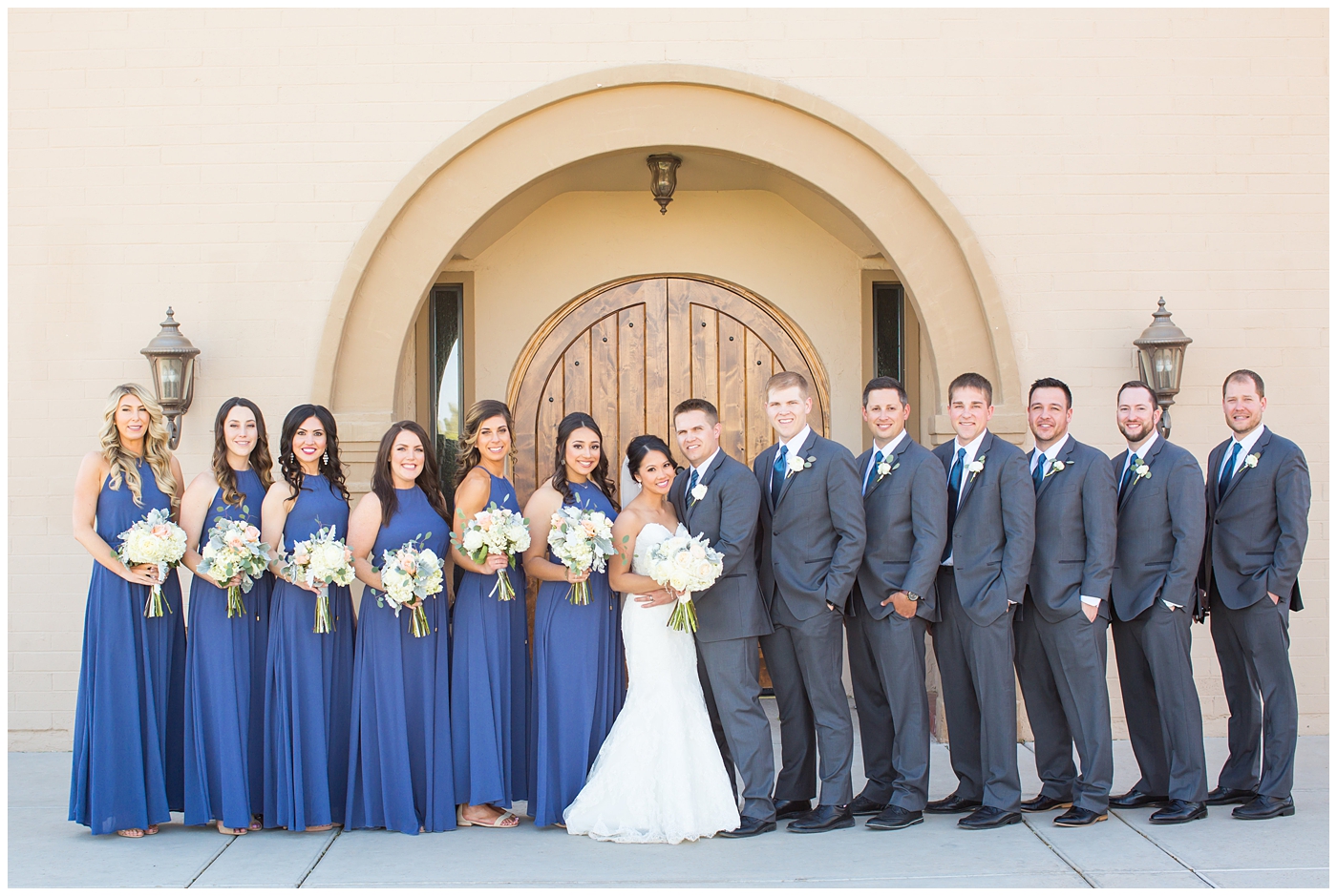 Bride in strapless maggie sorrento dress with soft white and pink roses, peonies, hydrangeas and greenery bouquet and groom in light gray suit with blue tie and white rose boutonniere with bridesmaids in blue maxi dresses and groomsmen in gray suits bridal party wedding day portrait 