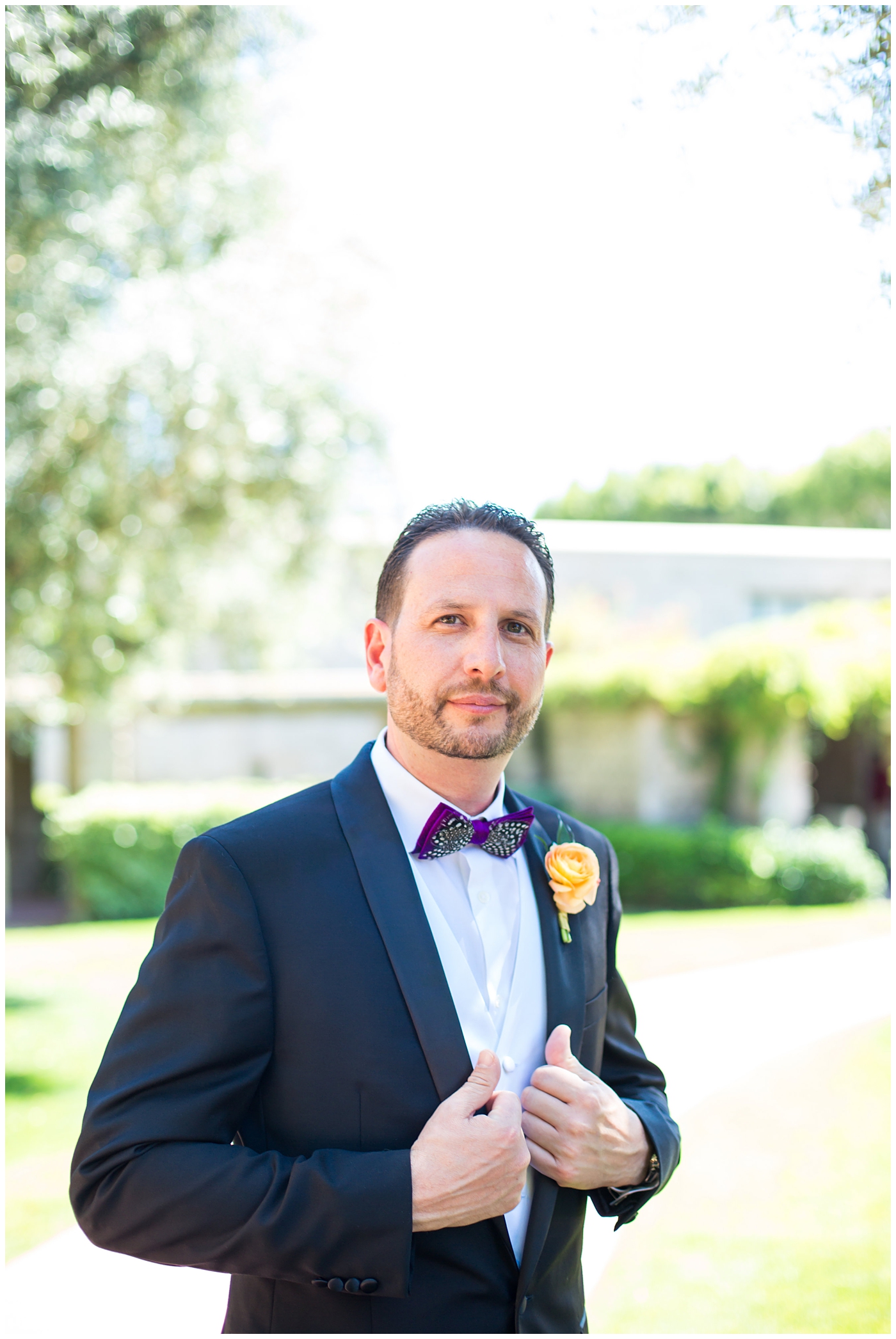 groom in black suit with purple bowtie with black and white feather and orange peony boutonniere wedding day portrait 