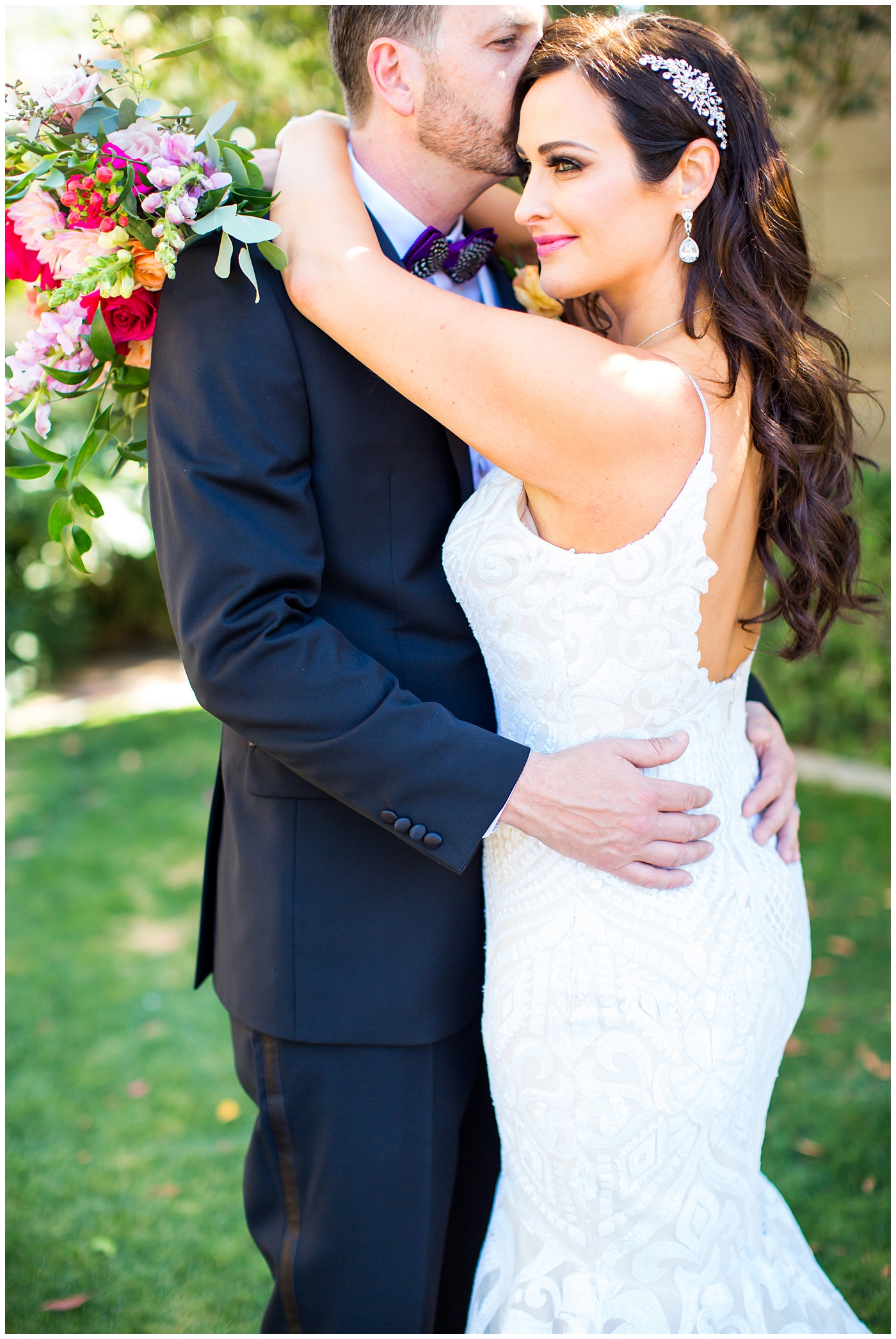 brunette bride in wedding dress with thin straps with bright color roses, dahlia, snap dragons loose bouquet with groom in black suit with purple bowtie with black and white feather with orange peony boutonniere wedding day portrait