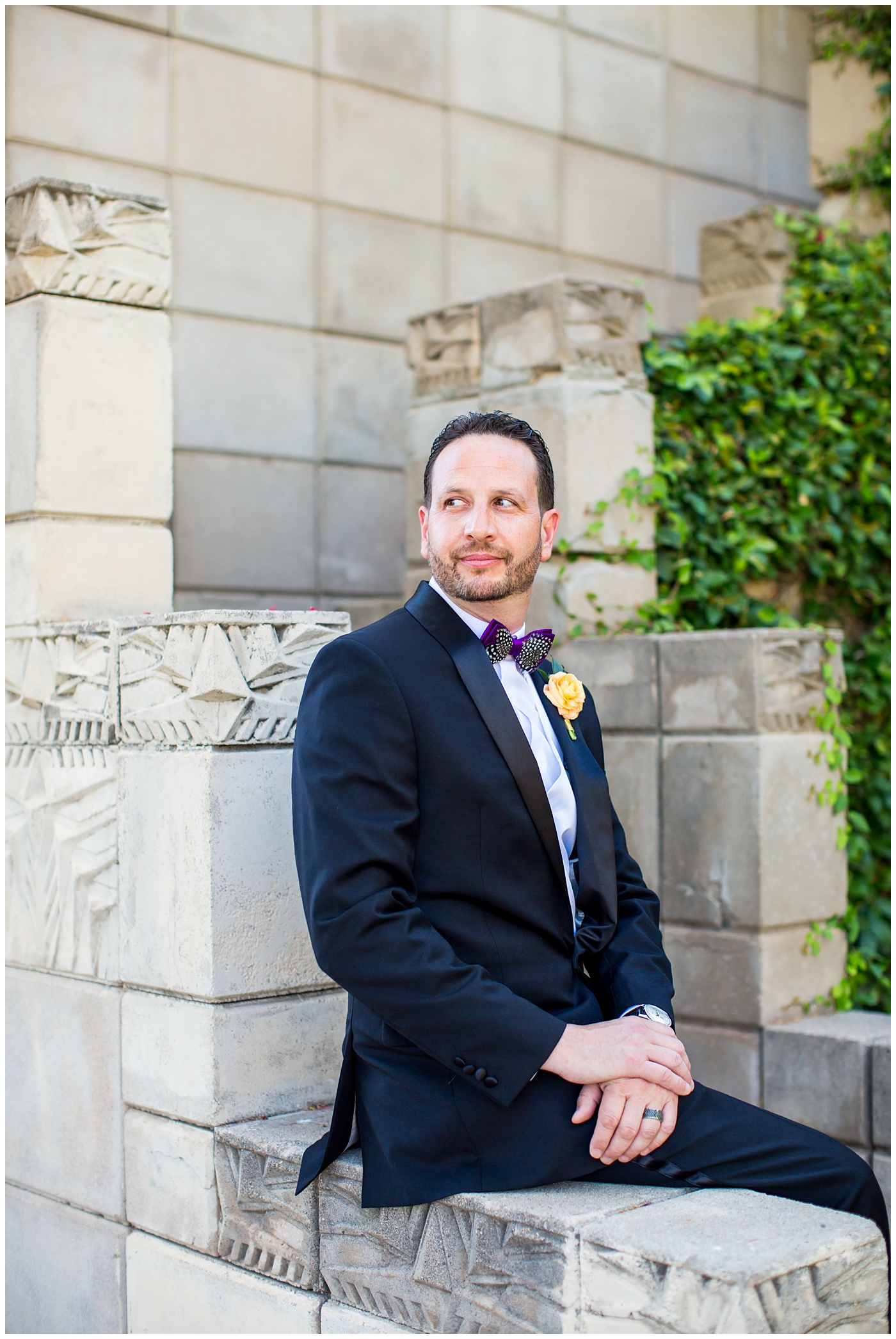 groom in black suit with purple bowtie with black and white feather with orange peony boutonniere wedding day portrait