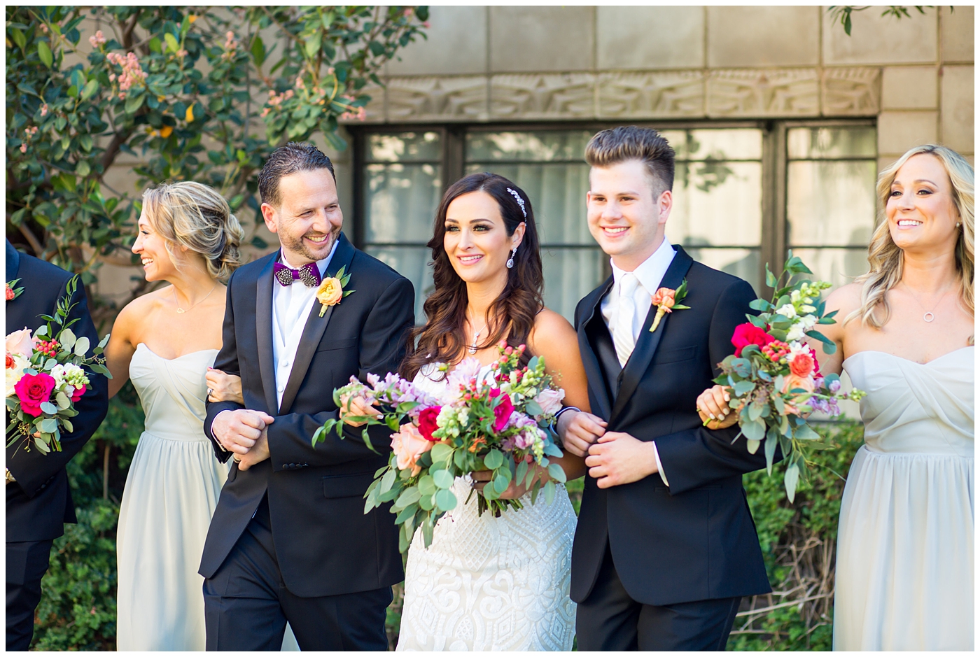 brunette bride in wedding dress with thin straps with bright color roses, dahlia, snap dragons loose bouquet with bridesmaids in champagne long dresses and groom in black suit with purple bowtie with black and white feather with orange peony boutonniere with groomsmen with white ties wedding day bridal party portrait