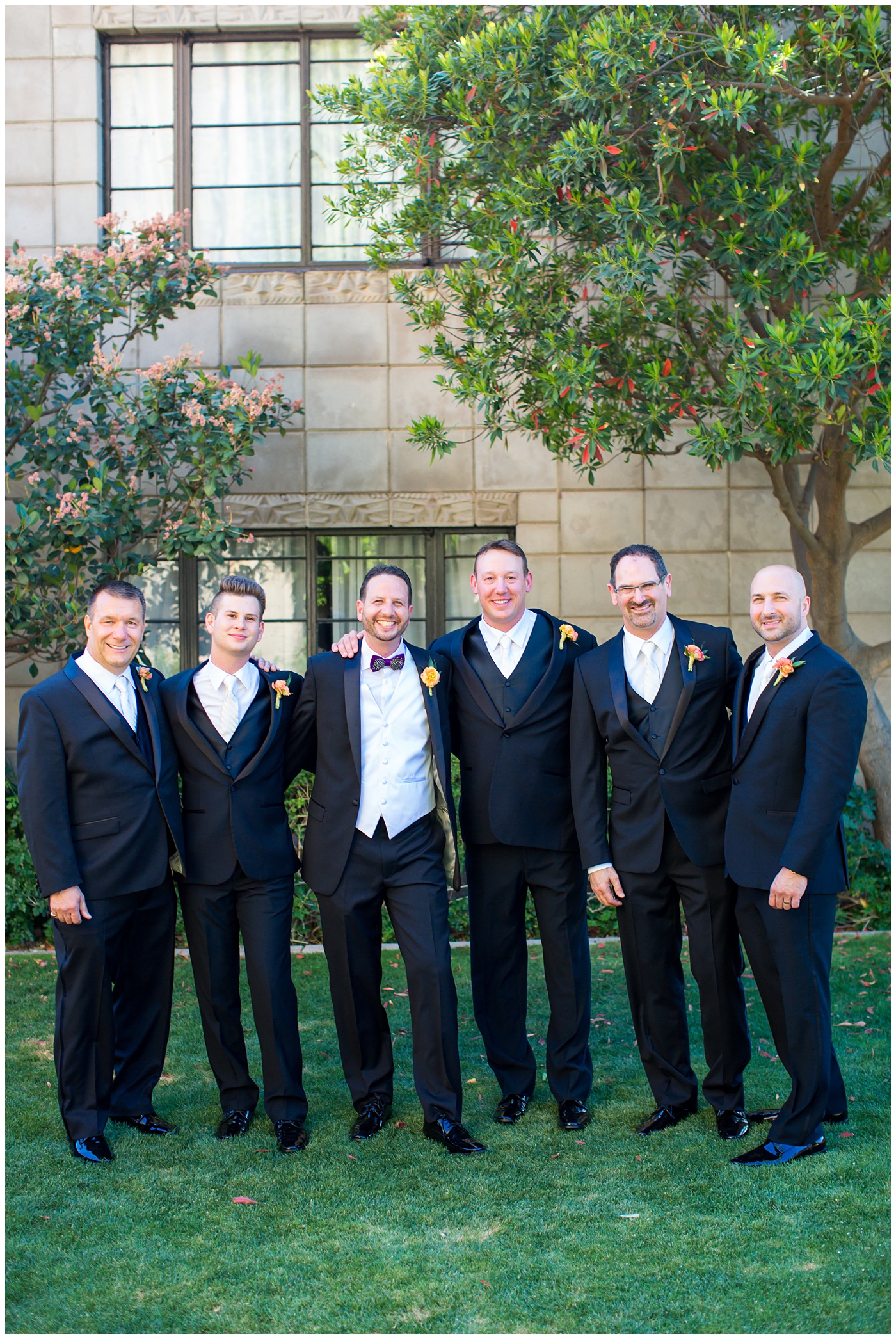 groom in black suit with purple bowtie with black and white feather with orange peony boutonniere with groomsmen with white ties wedding day bridal party portrait