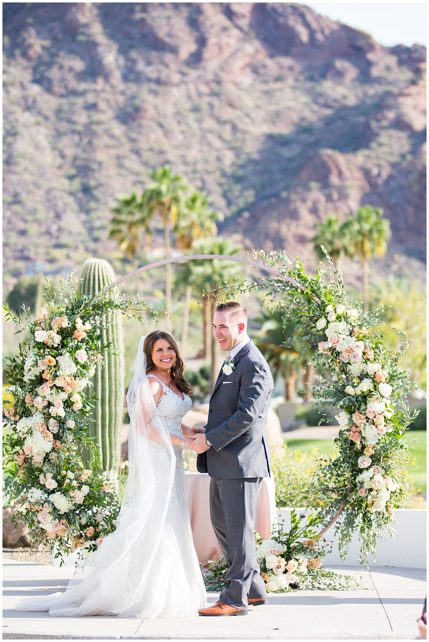 bride in wtoo watters wedding dress with lace straps with wildflower bridal bouquet with soft pink, blush, white, greenery flowers including roses, dahlias, snapdragons with groom in gray suit with pink tie and ranunculus boutonniere during their outdoor wedding ceremony with unique circle arch