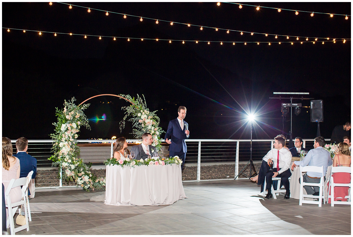 best man giving speech with unique circle arch for outdoor rooftop wedding reception with wildflowers in blush and white with greenery with twinkle lights stranded above