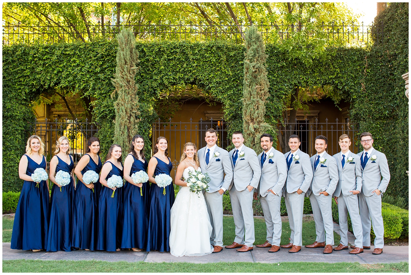 bride in strapless lace dress with white flower bouquet with roses and hydrangeas with bridesmaids in royal blue long dresses with blue hydrangeas bouquets with groom in grey suit with blue tie and white rose boutonniere and groomsmen in grey suits with royal blue vests and ties wedding party group shot 