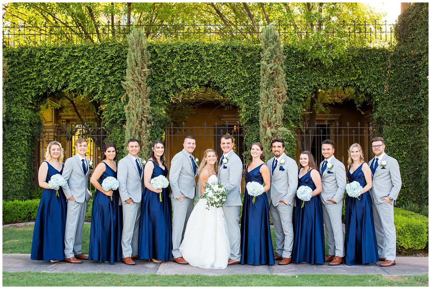 bride in strapless lace dress with white flower bouquet with roses and hydrangeas with bridesmaids in royal blue long dresses with blue hydrangeas bouquets with groom in grey suit with blue tie and white rose boutonniere and groomsmen in grey suits with royal blue vests and ties wedding party group shot 