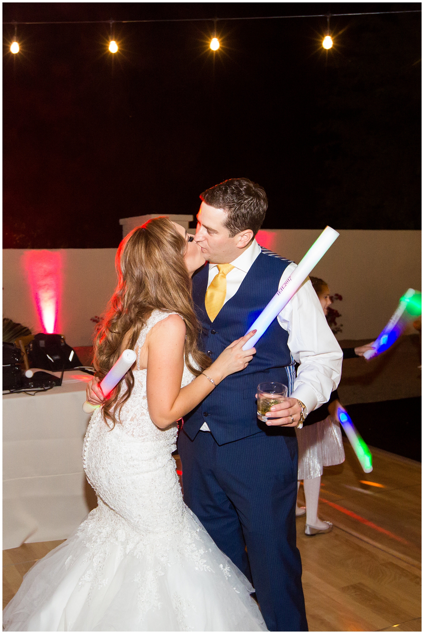 groom in blue suit with yellow tie and red rose boutonniere and bride in morilee wedding dress with red and white rose with light up rave sticks for wedding reception guests dancing at outdoor wedding reception with twinkle lights strung above