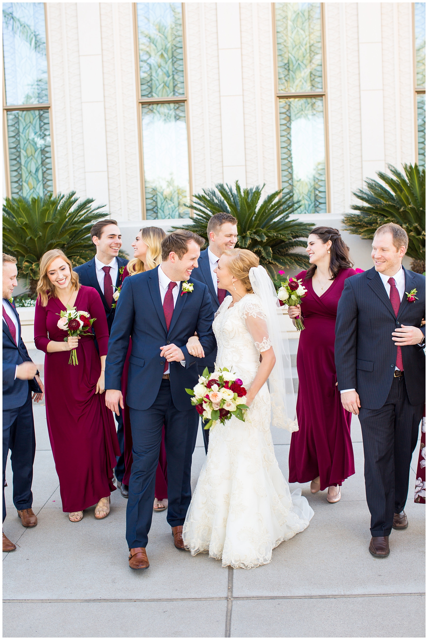 bride in sleeved lace dress with burgundy, white, and green wedding bouquet with bridesmaids in long sleeve burgundy dresses and groom in navy suit with burgundy tie with groomsmen wedding party portrait at Gilbert LDS temple