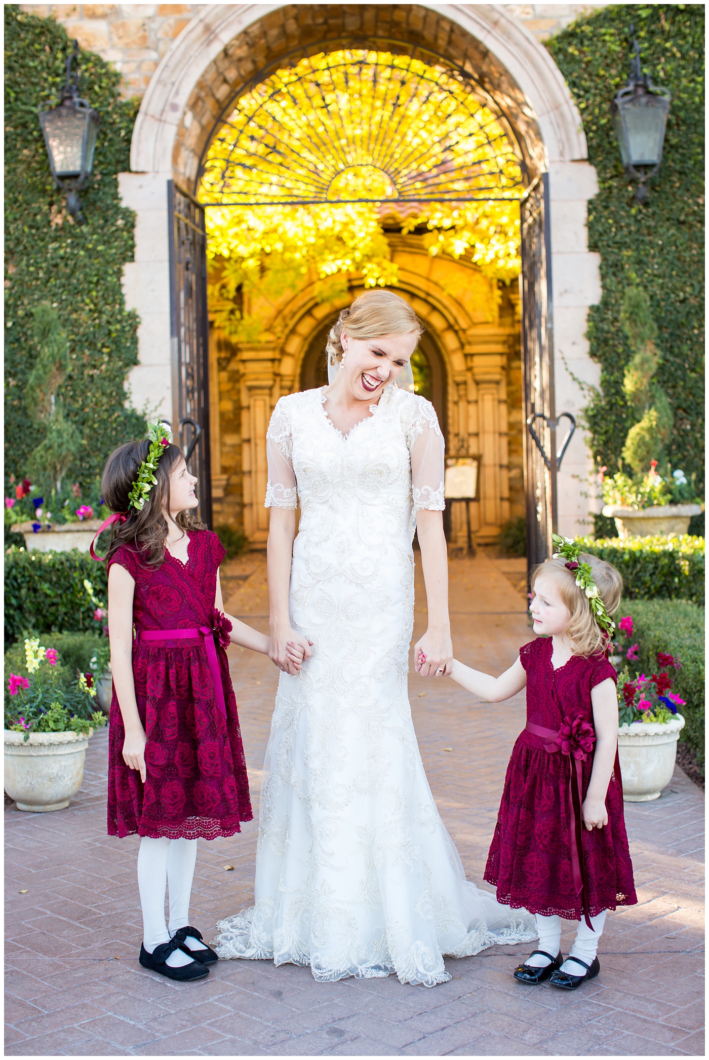 bride in sleeved lace dress with burgundy, white, and green wedding bouquet wedding day portrait with flower girls with floral head bands and burgundy dresses at Villa Siena