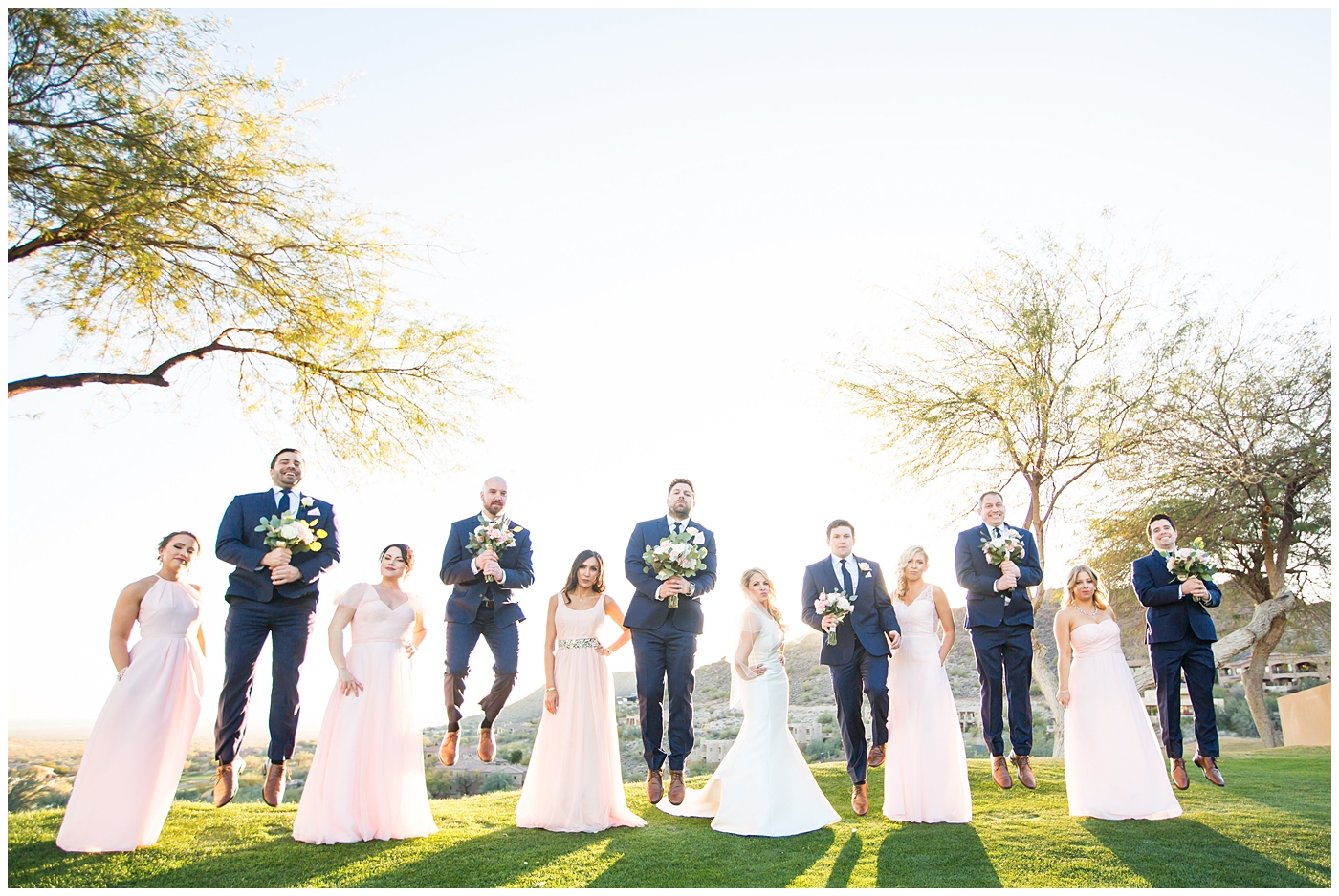 gorgeous bride with side swept hair in racerback pronovias dress and blush pink, white rose and eucalyptus greenery wedding bouquet with groom in navy suit and tie with bridal party bridesmaids in blush pink dresses and groomsmen in navy blue suits on wedding day