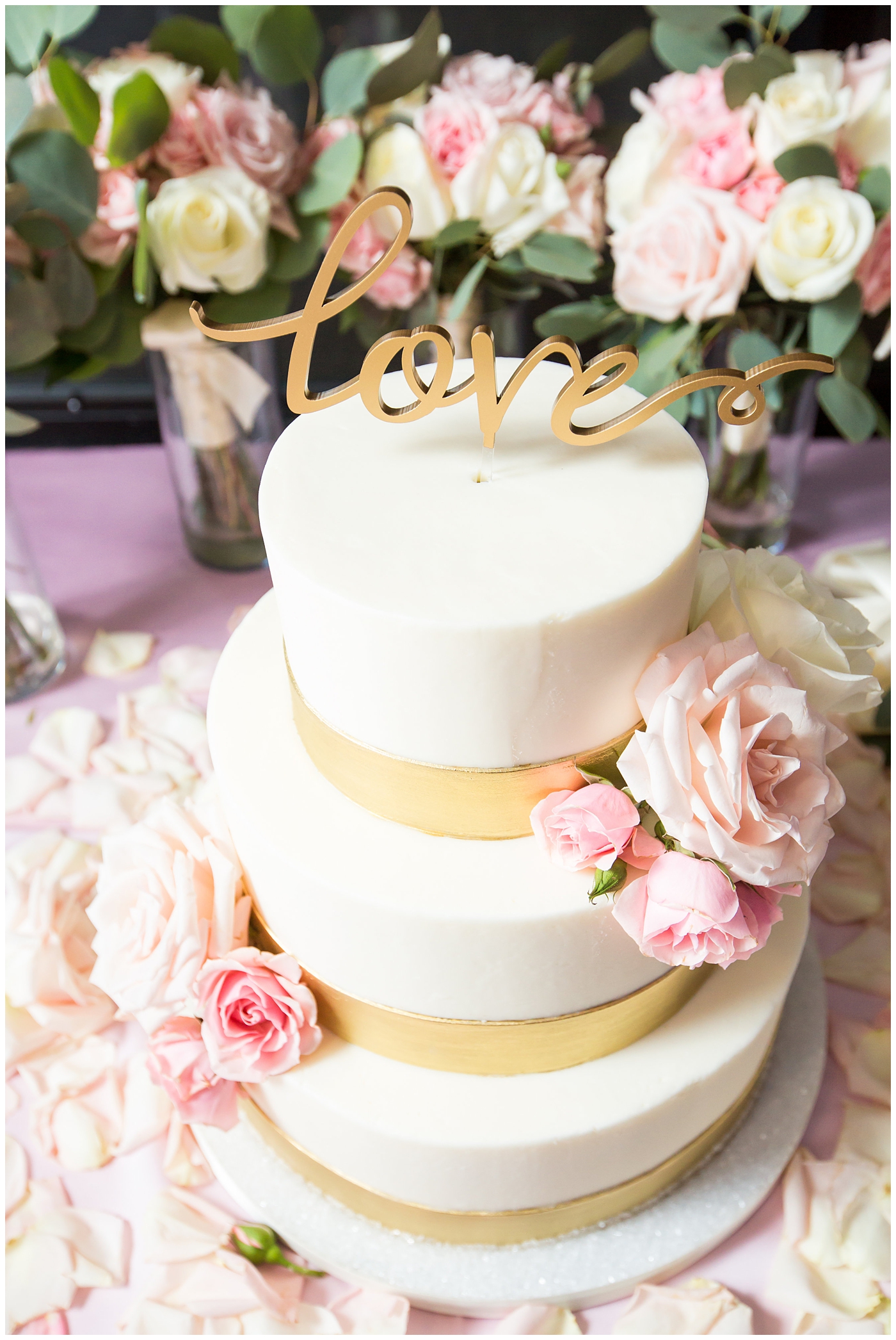 white smooth three tier wedding cake with gold ribbon and gold love cutout wedding cake topper at reception
